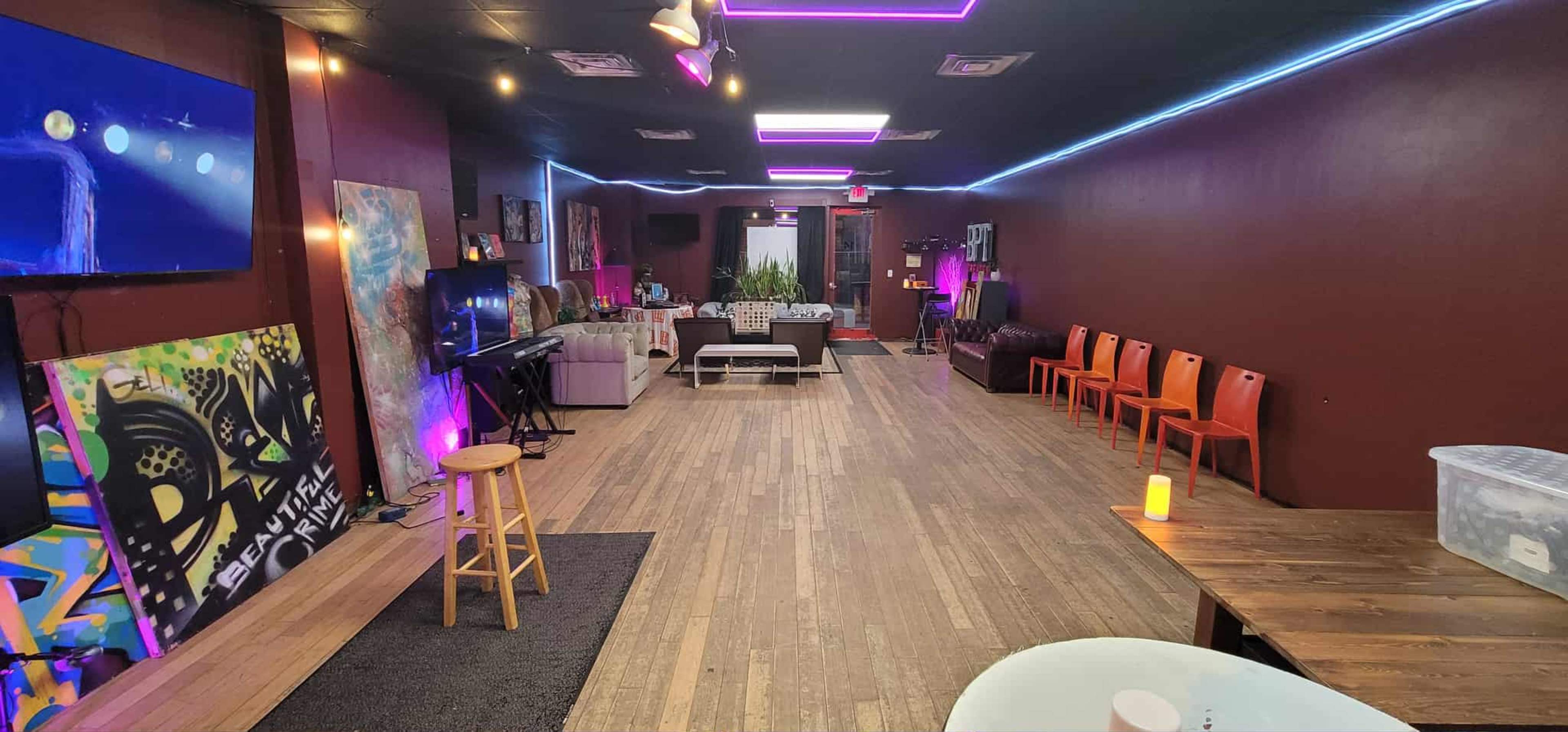 The image shows an interior space with a long wooden floor, vibrant artwork on the walls, a seating area with orange chairs, and a small stage setup at one end.