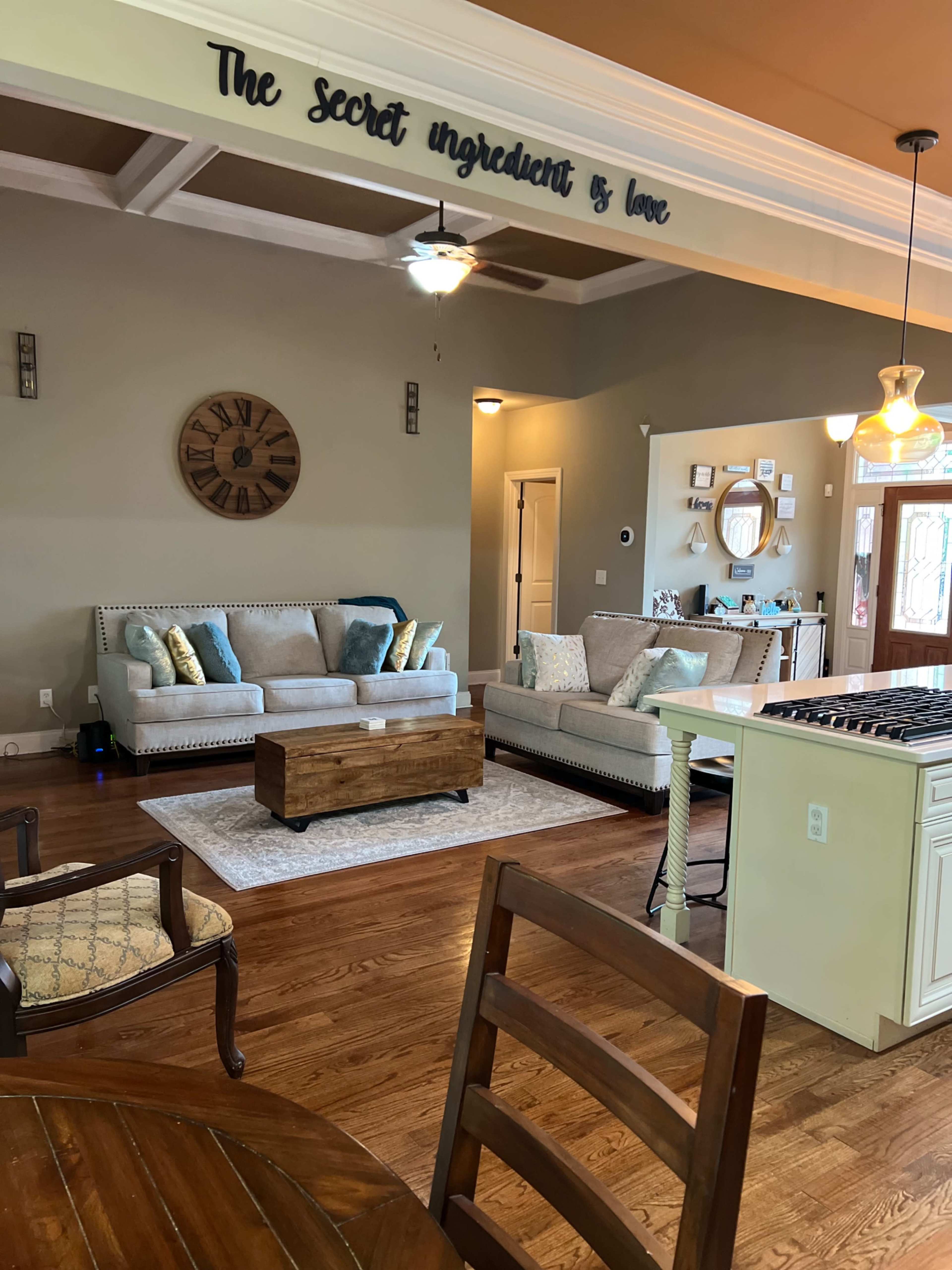 A living room and kitchen area featuring a beige sofa, wooden coffee table, and wall decor with the phrase "The secret ingredient is love."