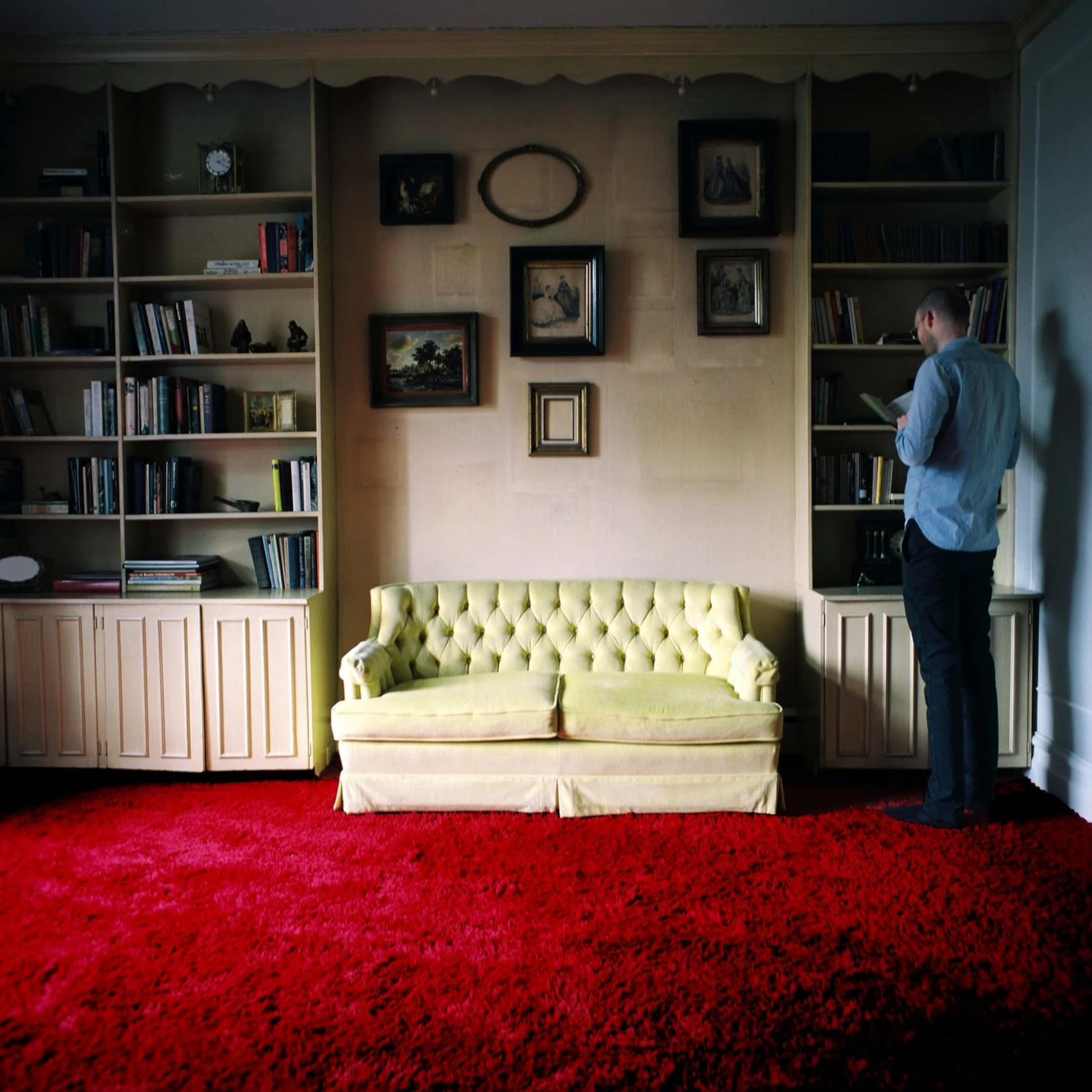 A person stands next to a bookshelf in a room featuring a yellow tufted sofa and a red carpet.
