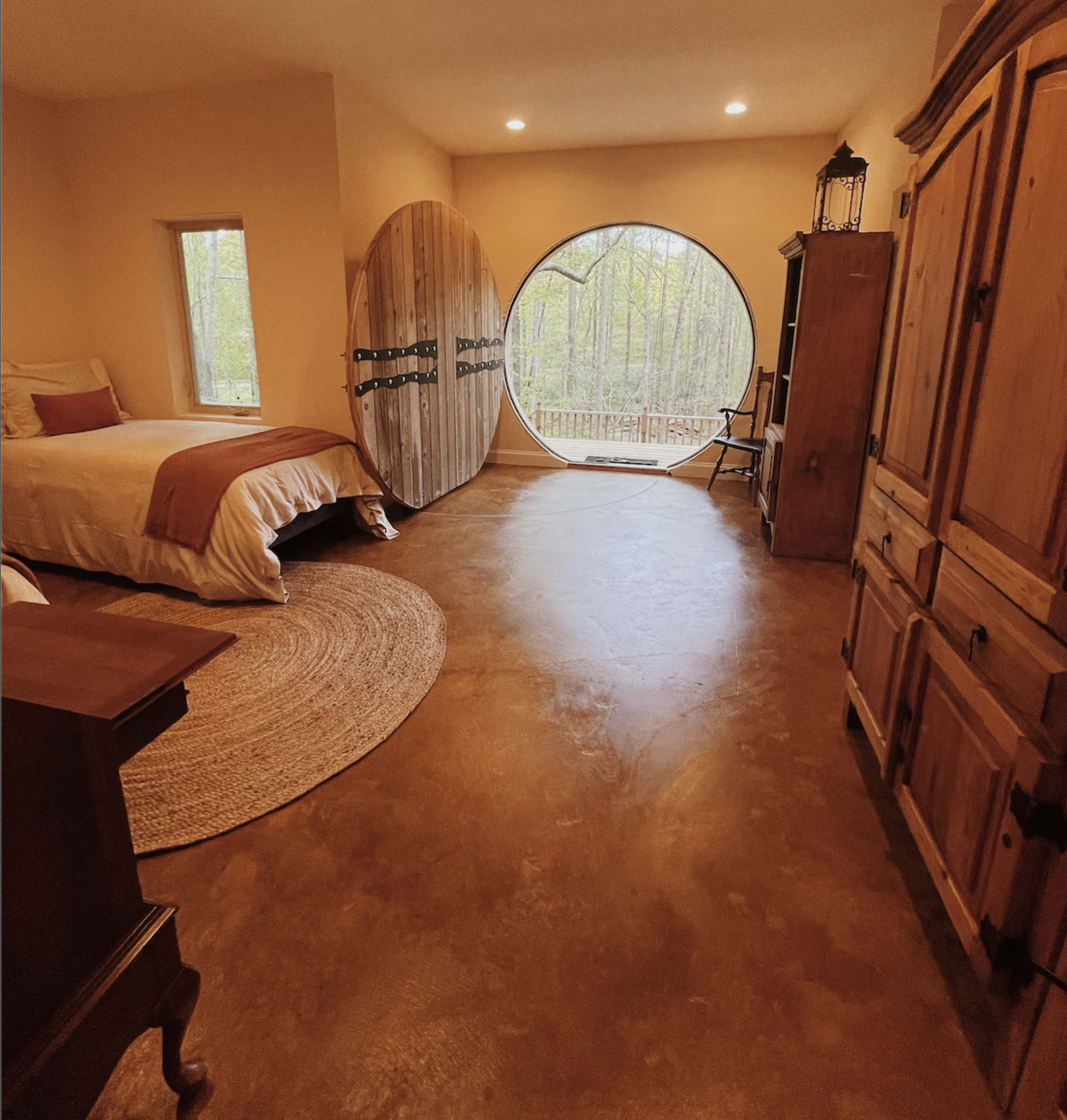 A bedroom with a round window facing a wooded area, featuring a wooden door, a bed, and rustic furniture.