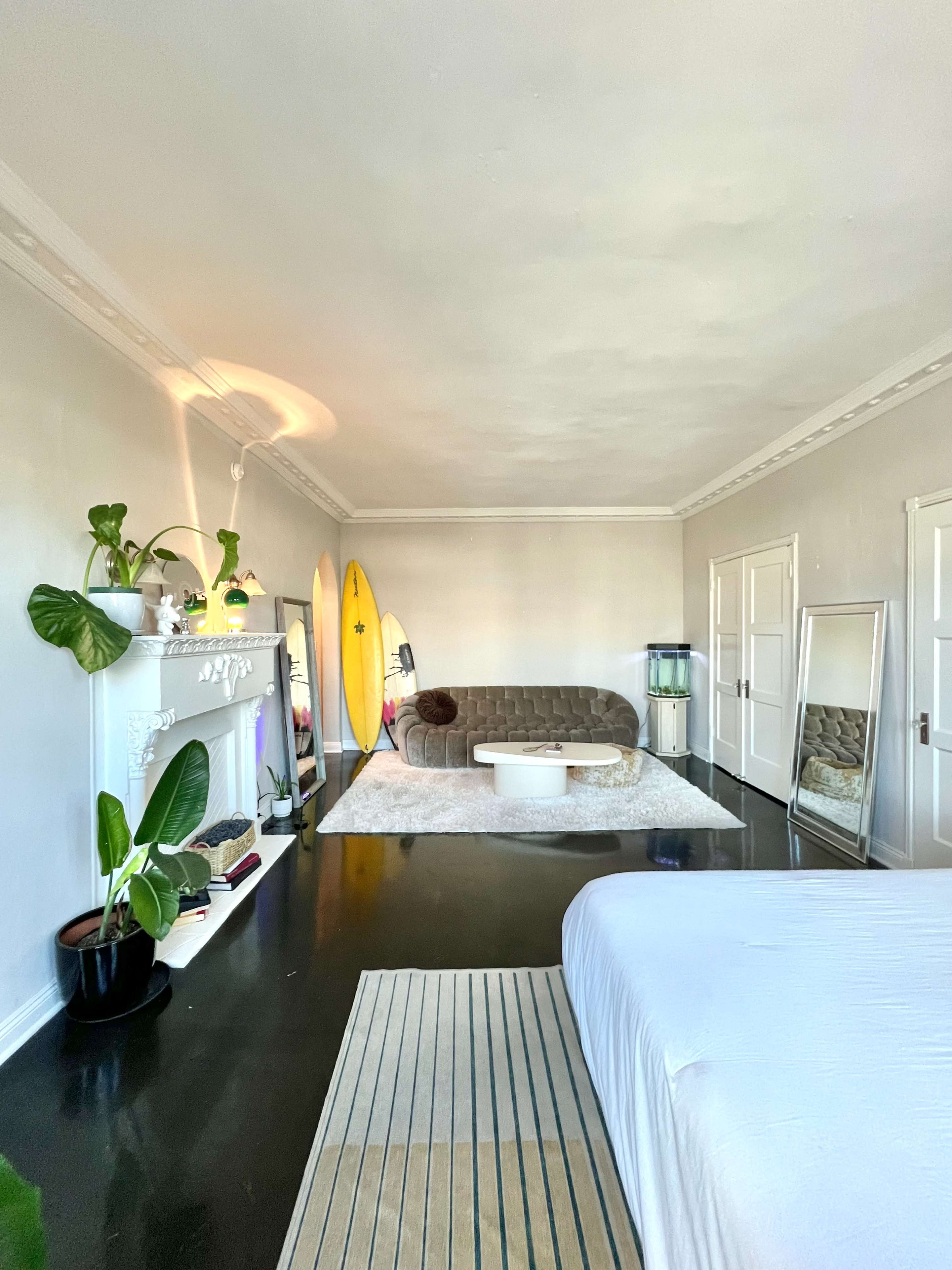 The room features a living space with a sofa, a fireplace, a rug, decorative plants, and surfboards leaning against the wall.