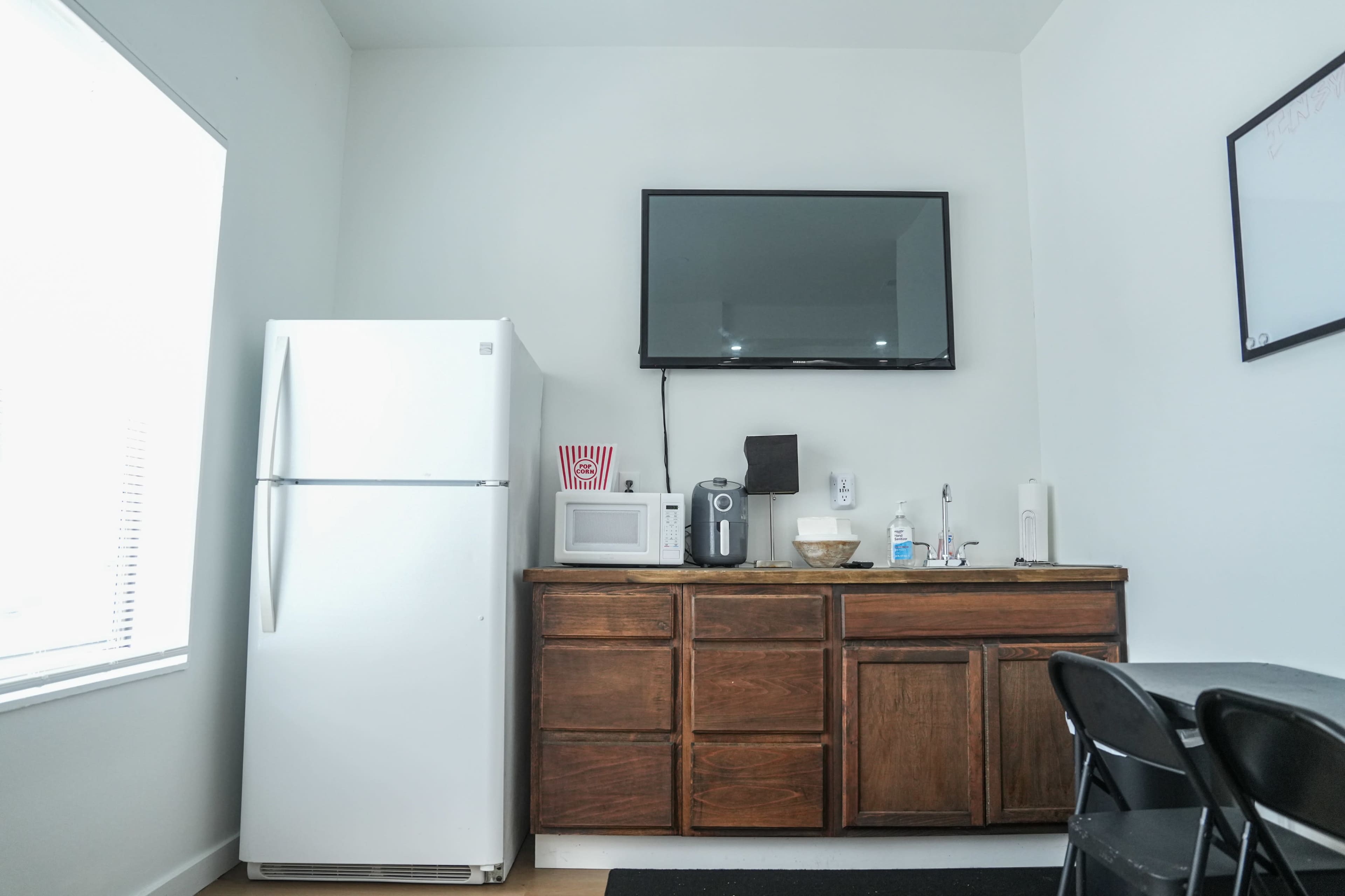 The image shows a small kitchen area featuring a white refrigerator, microwave, coffee maker, and a wooden cabinet with multiple drawers, alongside a wall-mounted television.