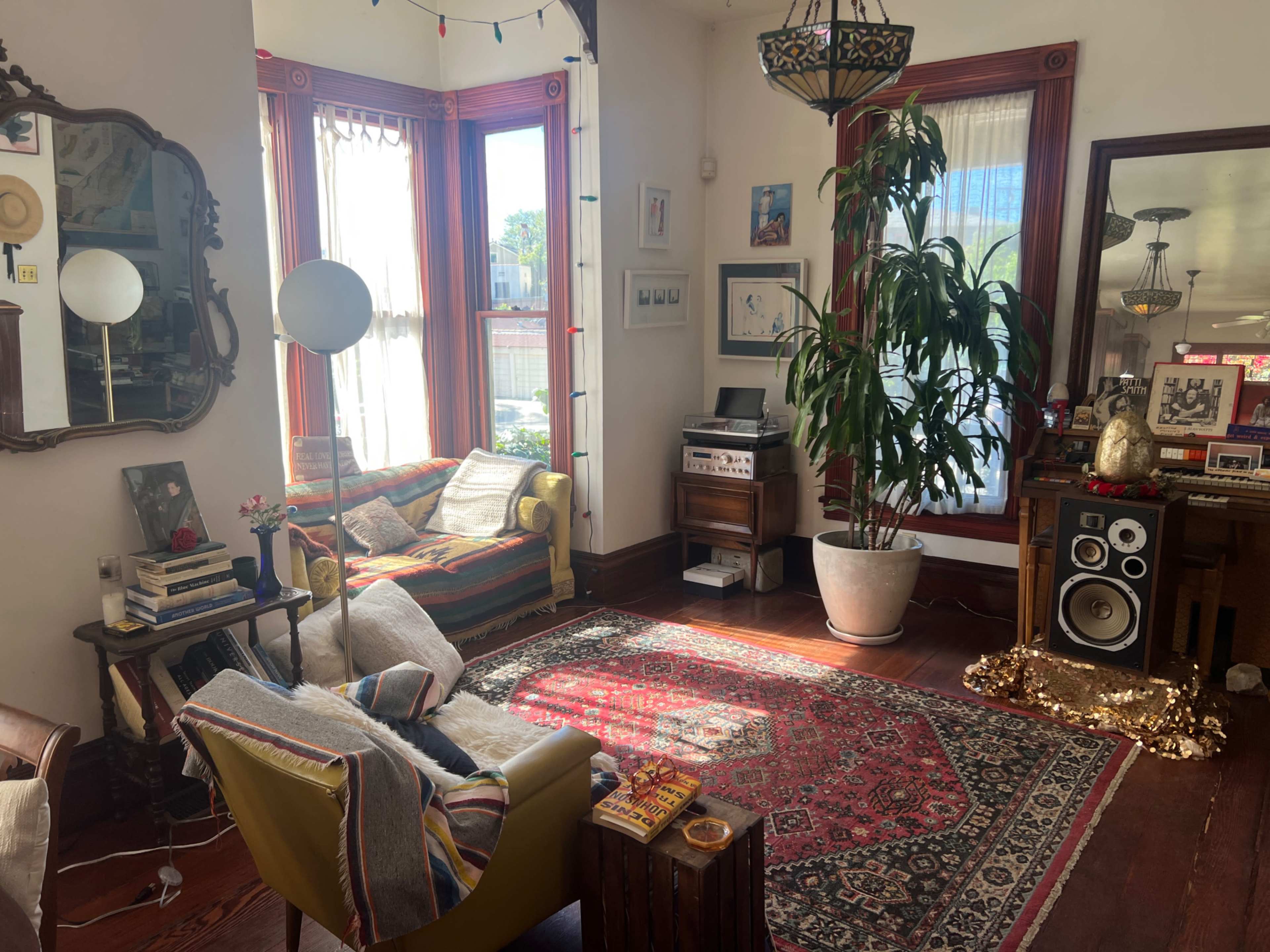 The image shows a bright living room featuring a colorful couch, a large potted plant, a vintage record player, and patterned rugs on wooden flooring.