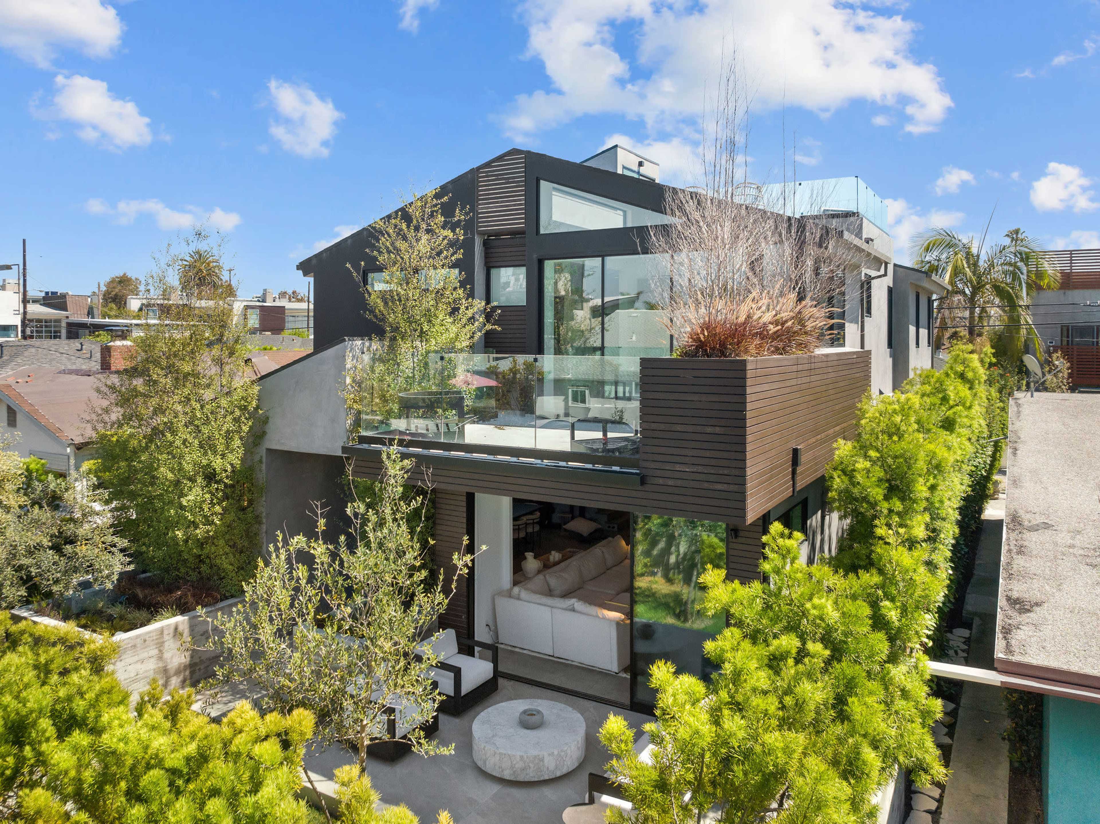 A modern two-story house features large glass windows and a spacious balcony surrounded by greenery.