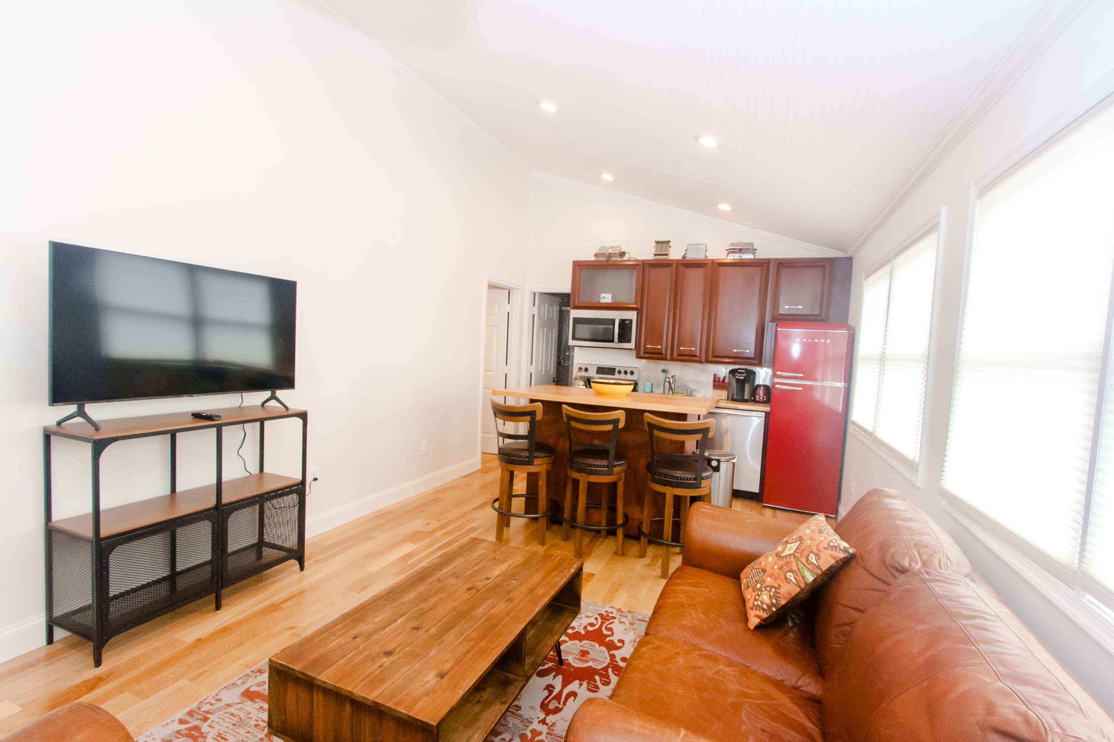 A bright living area with a leather couch, a wooden coffee table, a television on a stand, and an open kitchen featuring a red refrigerator and bar seating.