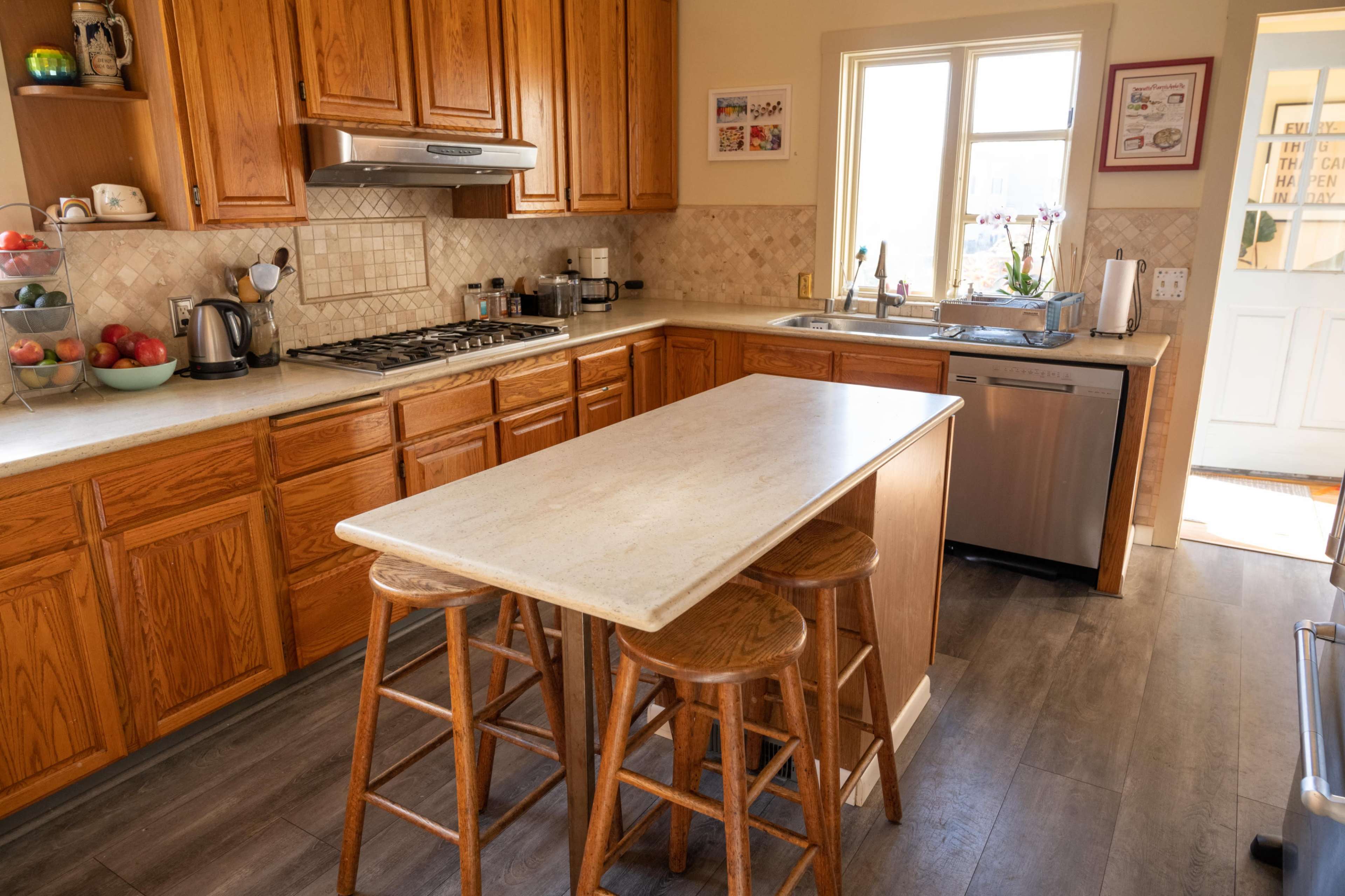 A kitchen with wooden cabinets, a countertop island with three stools, and a window facing an outdoor entrance.
