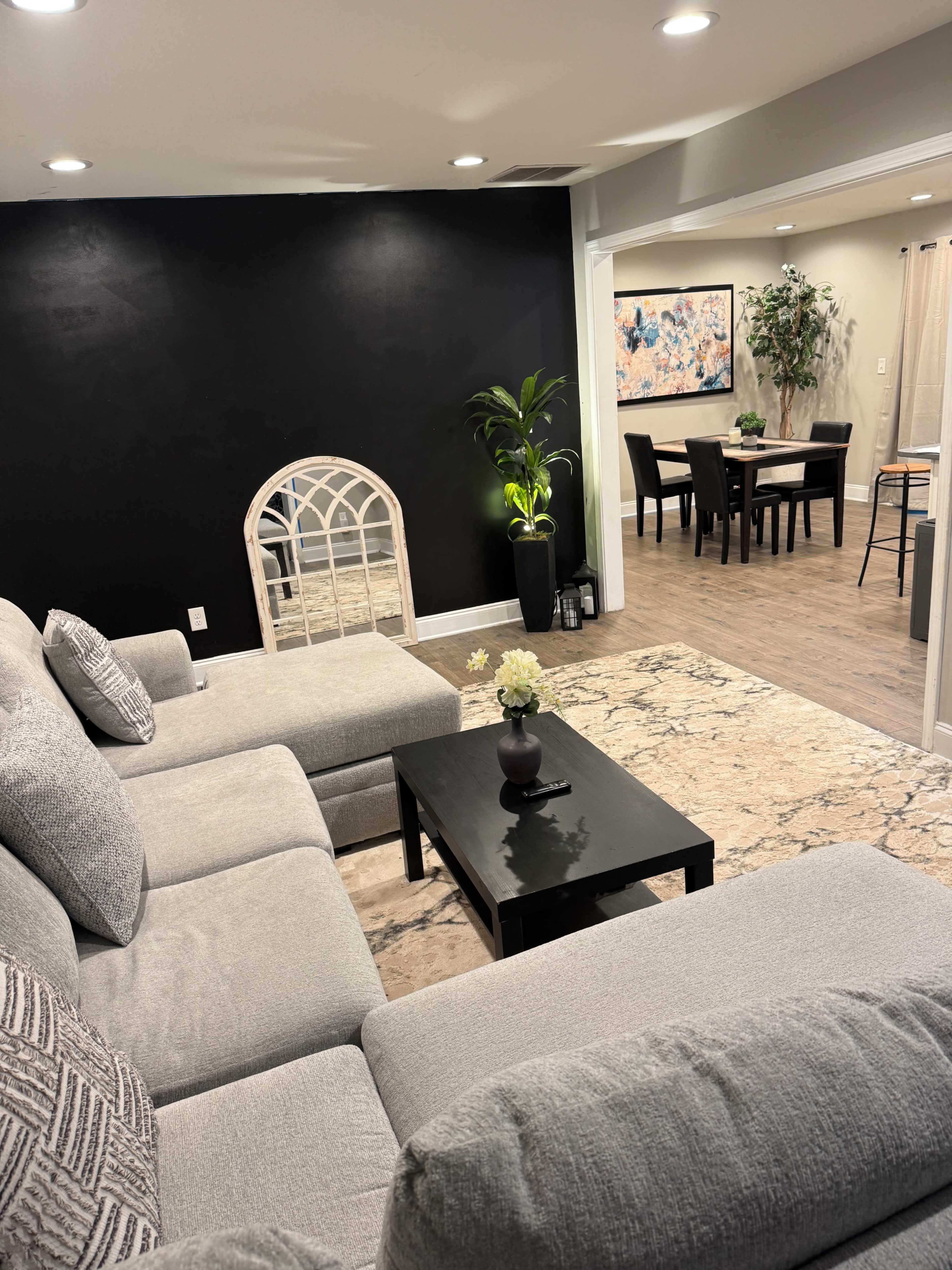 The image shows a modern living room with a gray sectional sofa, a black coffee table with a flower centerpiece, and a wall featuring a large mirror-like arch, adjacent to a dining area with a table and chairs.