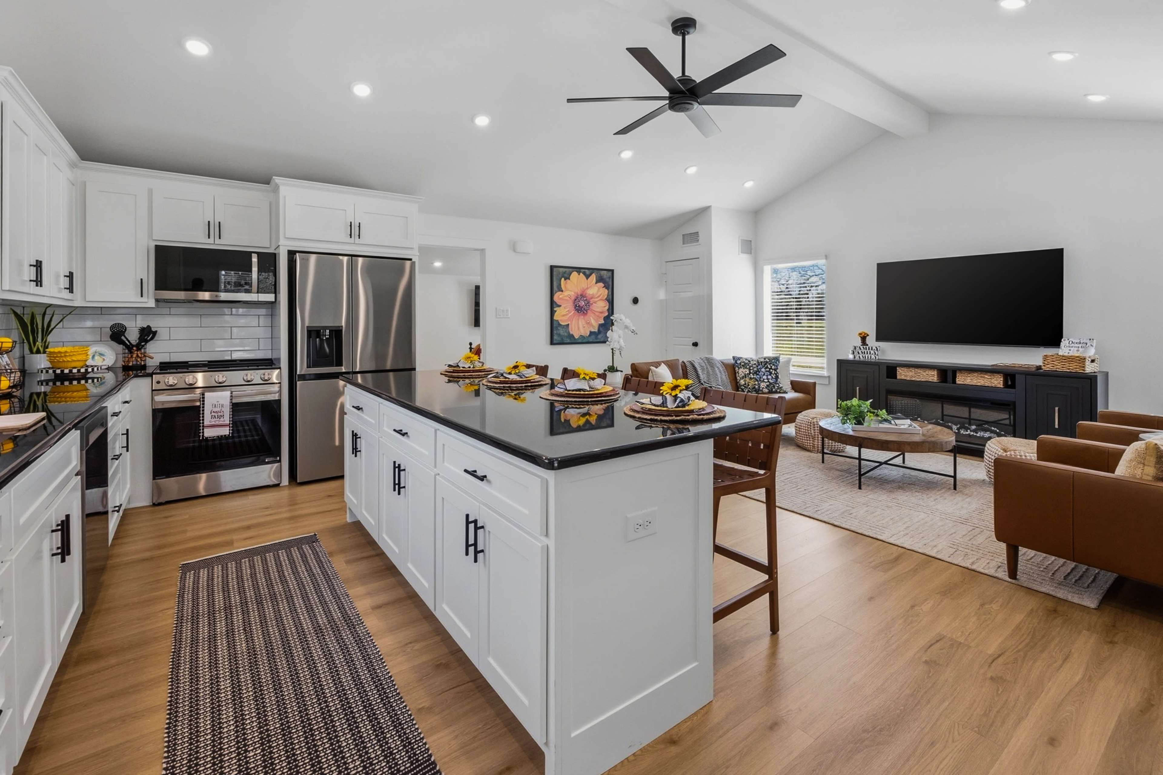 The image shows a modern kitchen with white cabinets, a large central island, and stainless steel appliances, leading into a living area with a television and seating.