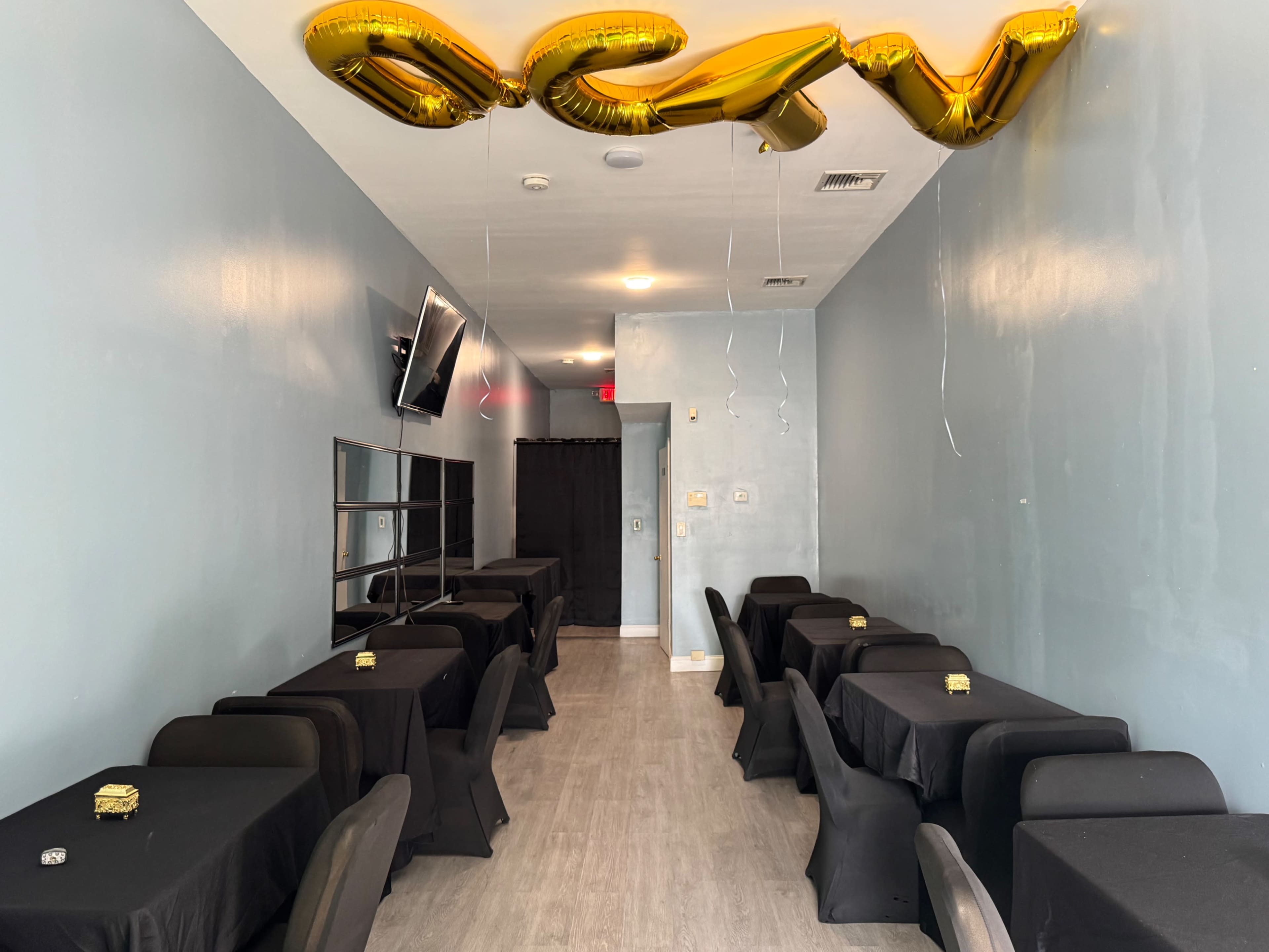 The image shows a narrow room with black tables covered by dark tablecloths, a television mounted on one wall, and gold balloons spelling "OPEN" hanging from the ceiling.