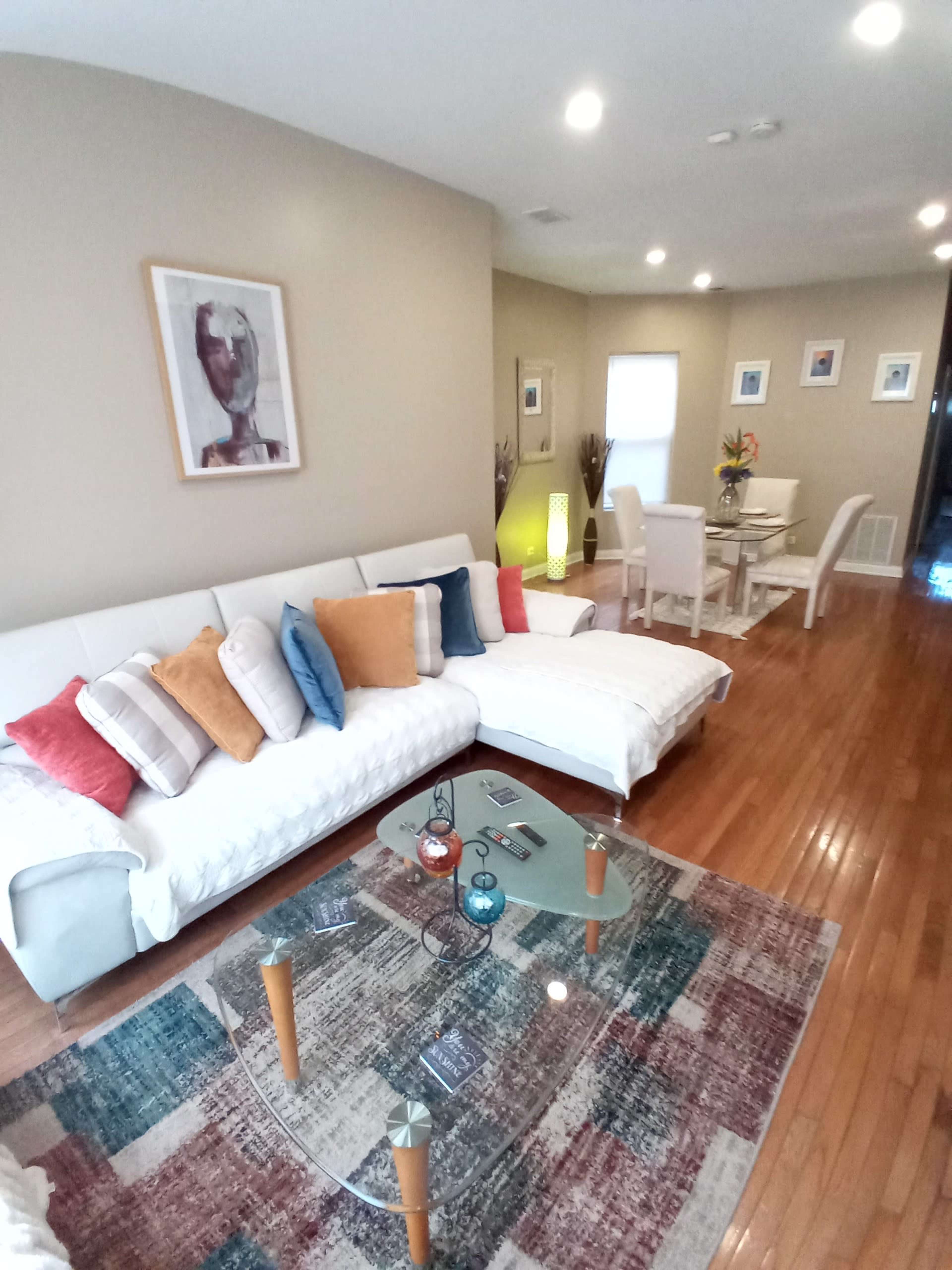 A living room with a white sectional sofa, a glass coffee table, and a dining area visible in the background.