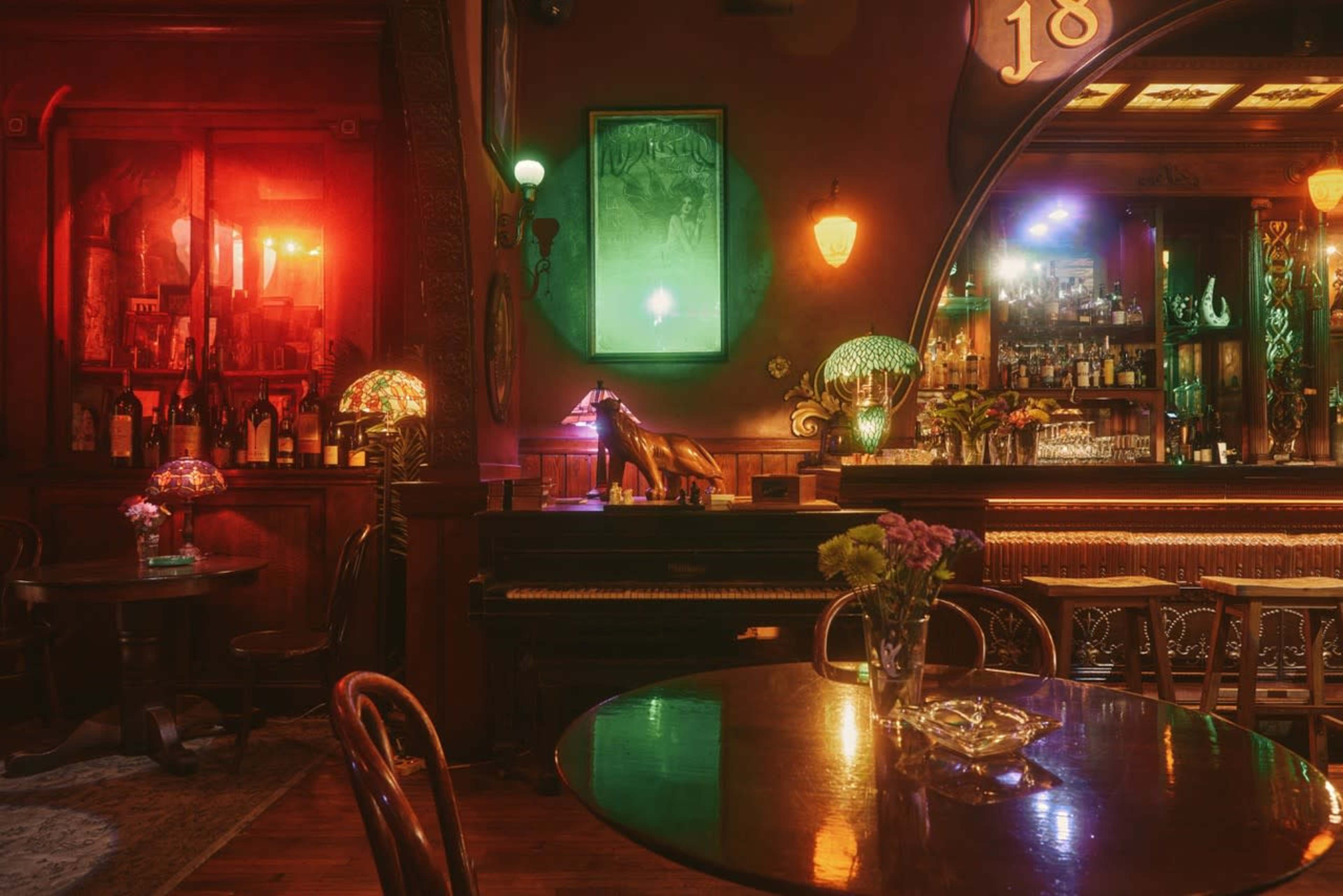 The image shows a dimly lit bar with vintage decor, featuring a piano, tables with flowers, and a well-stocked bar illuminated by colorful lights.