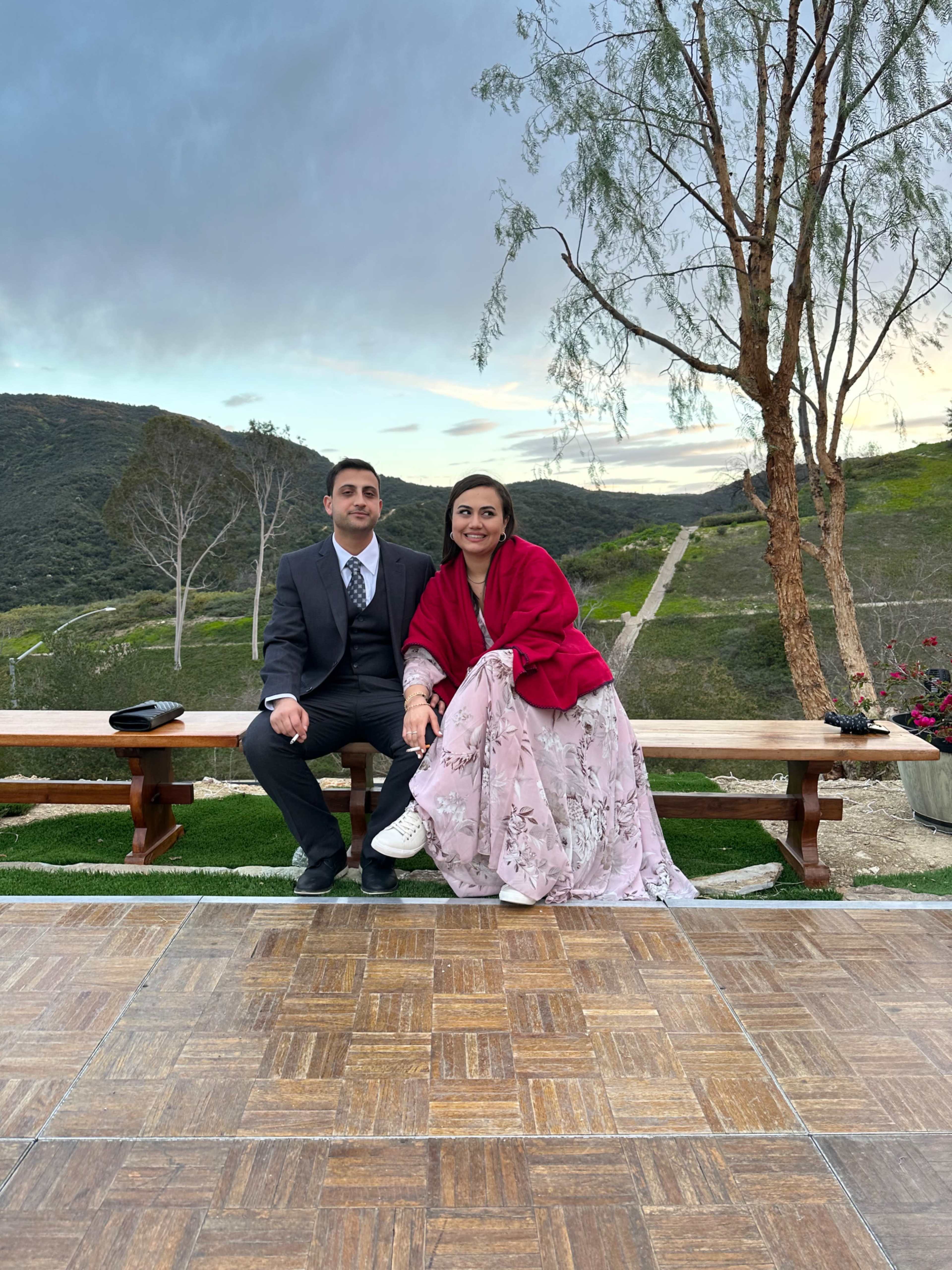 A man in a suit and a woman in a pink dress and red shawl sit on a wooden bench with a scenic hillside in the background.