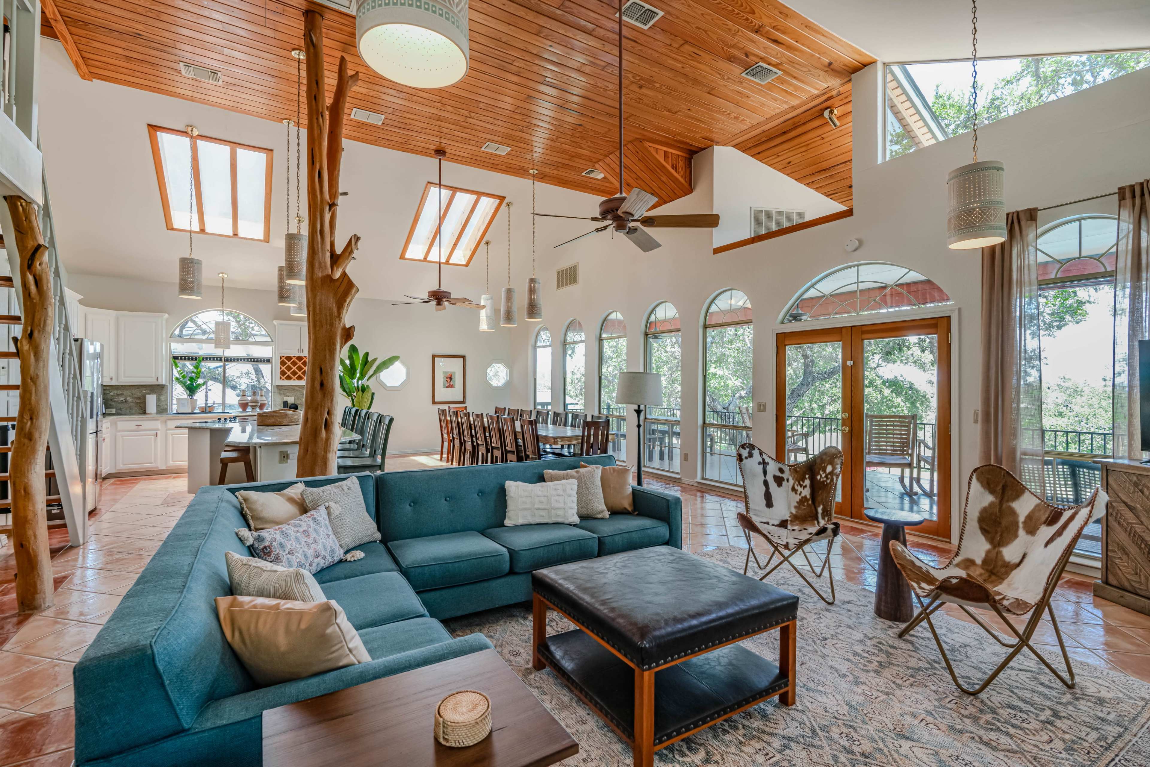 The image depicts a spacious living area with a large sectional sofa, wooden ceiling beams, and large windows that provide ample natural light.