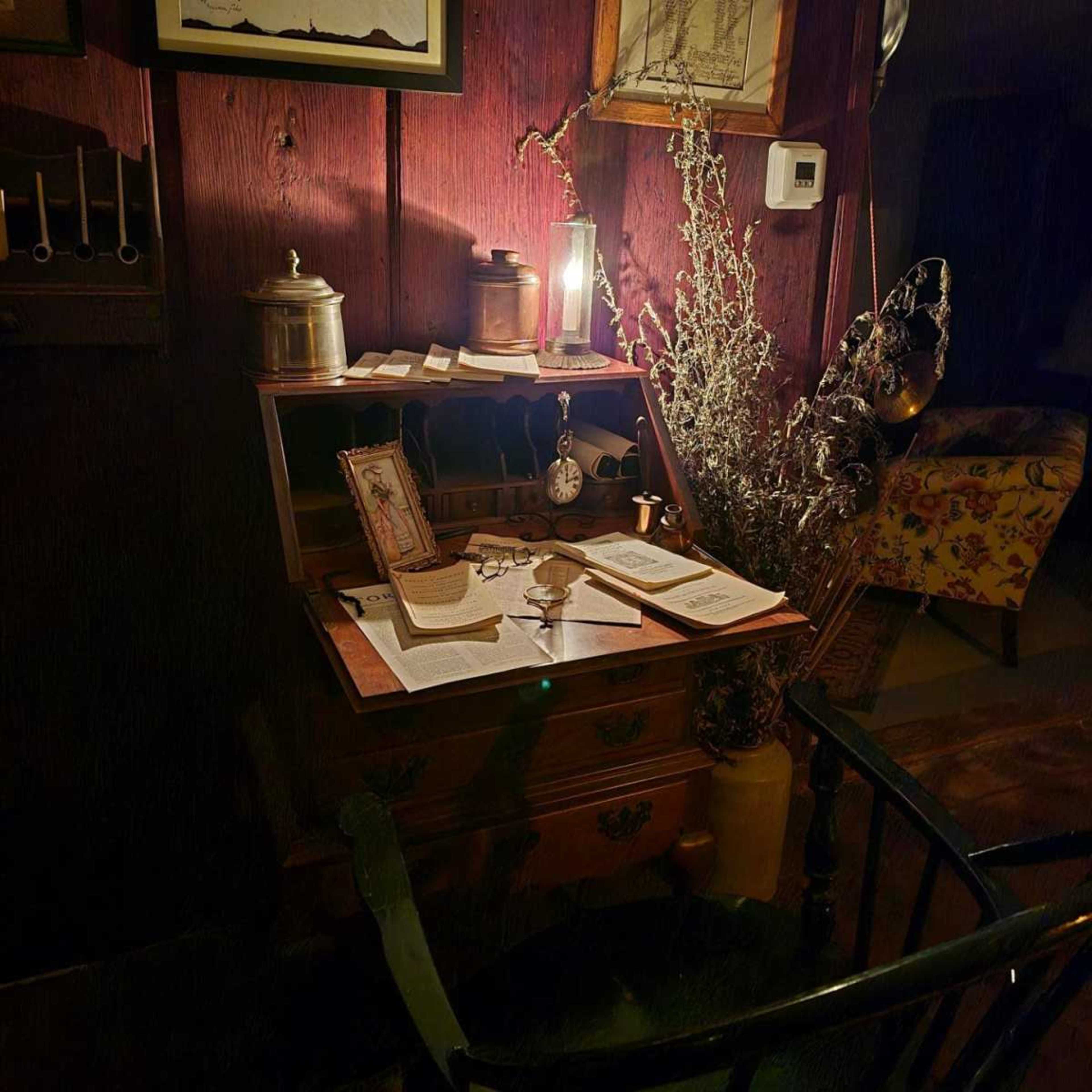 The image depicts an antique writing desk with a lamp, various papers, a clock, and a decorative arrangement of dried plants in a dimly lit room.