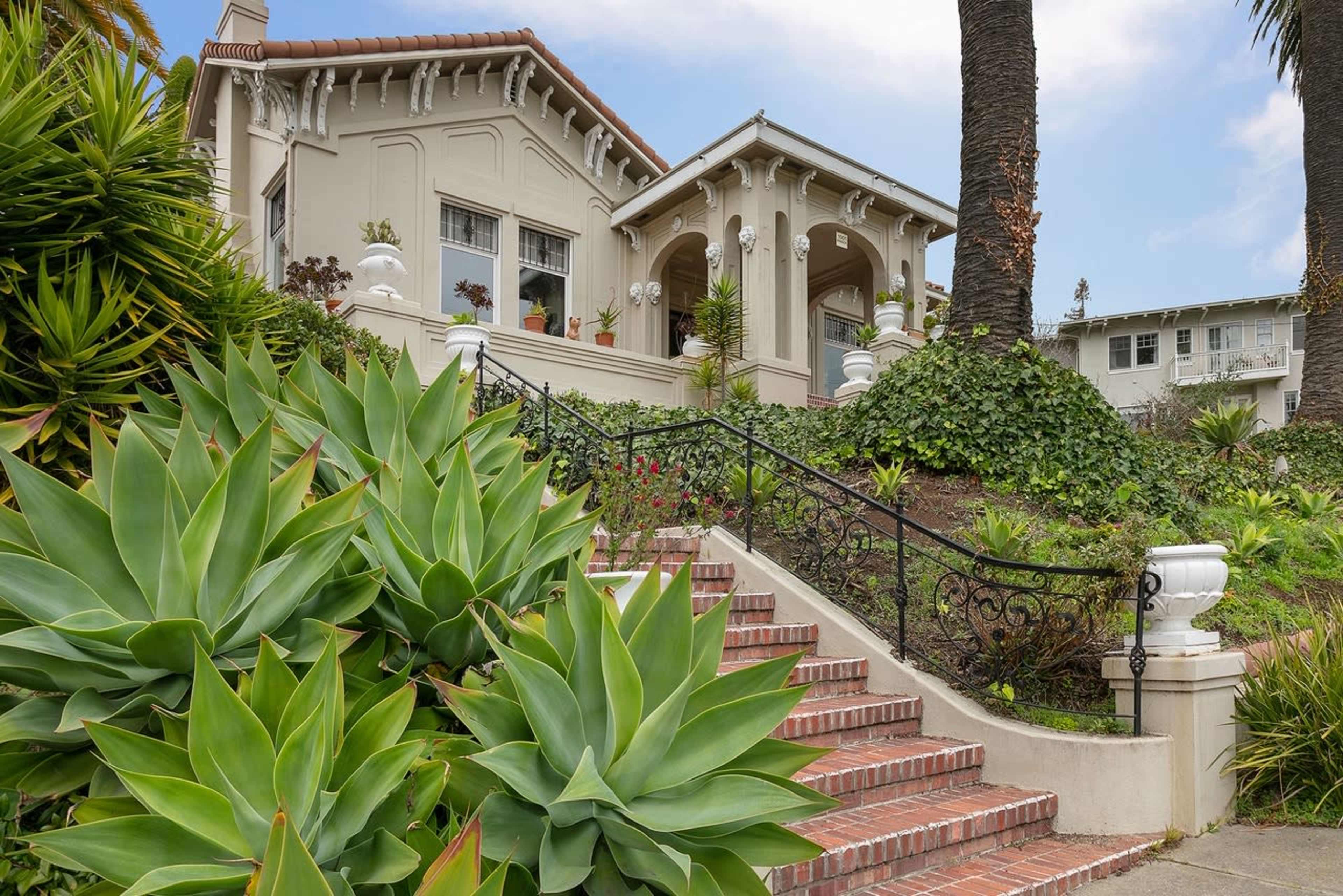 A large, two-story house with decorative arches and a staircase surrounded by lush greenery and palm trees.