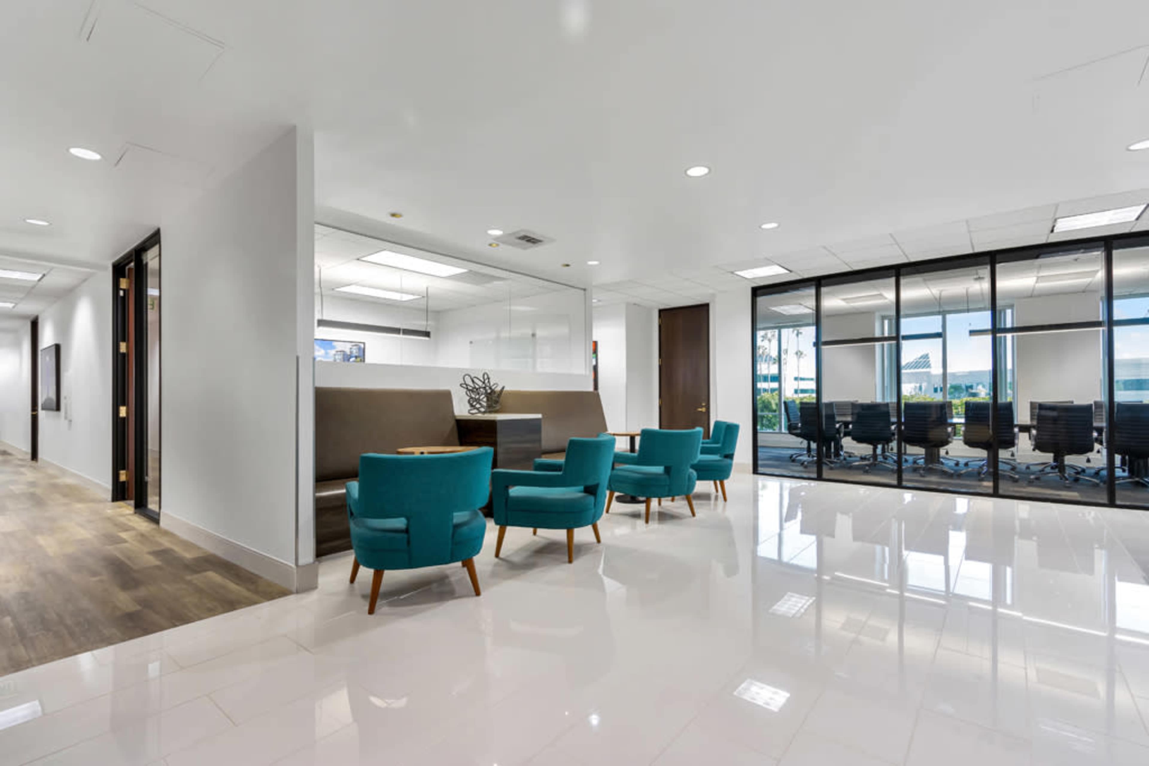 The image shows a modern office lobby featuring a seating area with teal chairs and a glass-walled conference room in the background.