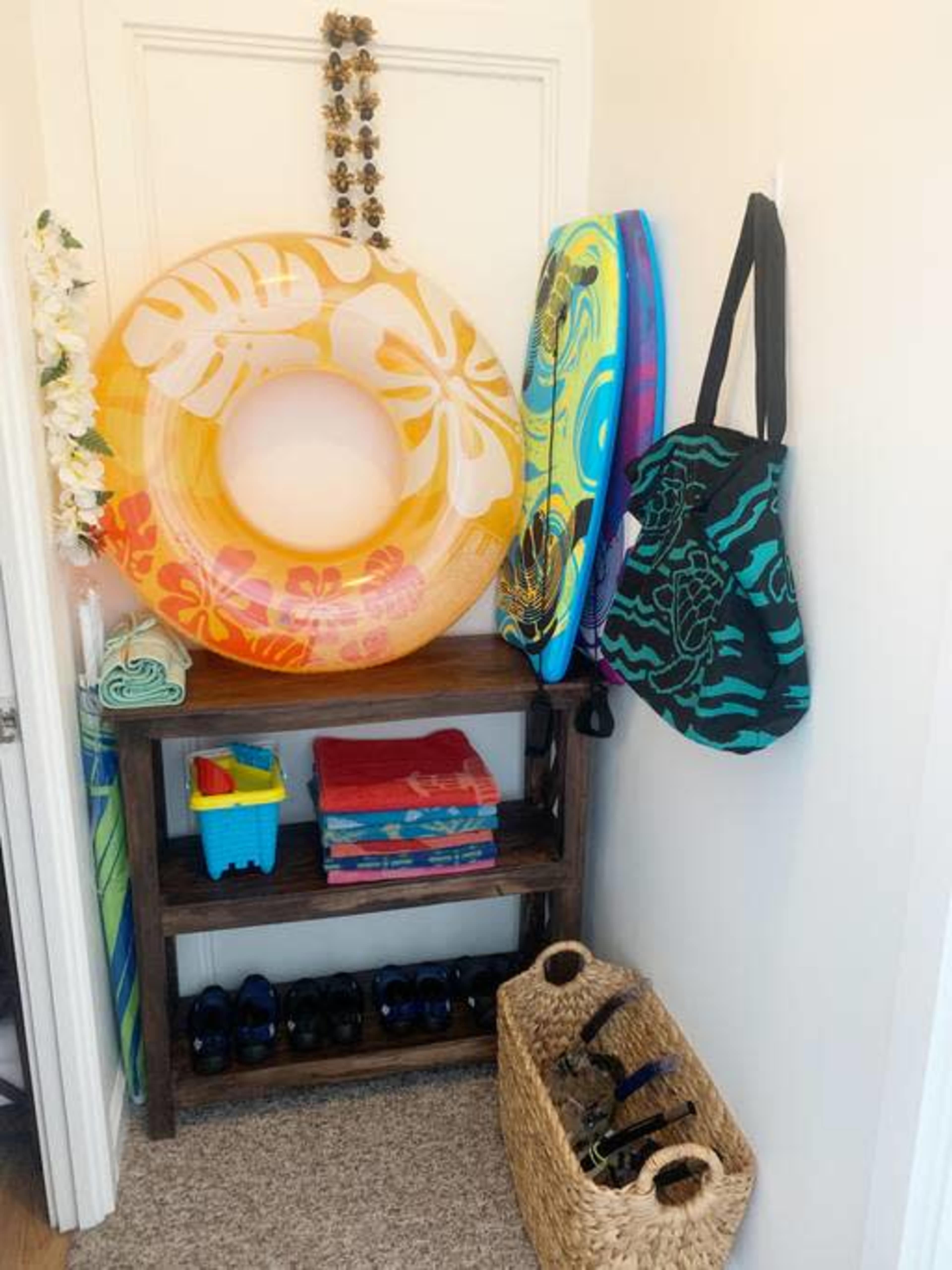 A corner with a wooden shelf holding towels and a blue basket, alongside an inflatable float, two colorful boards, a black tote bag, and several pairs of shoes.