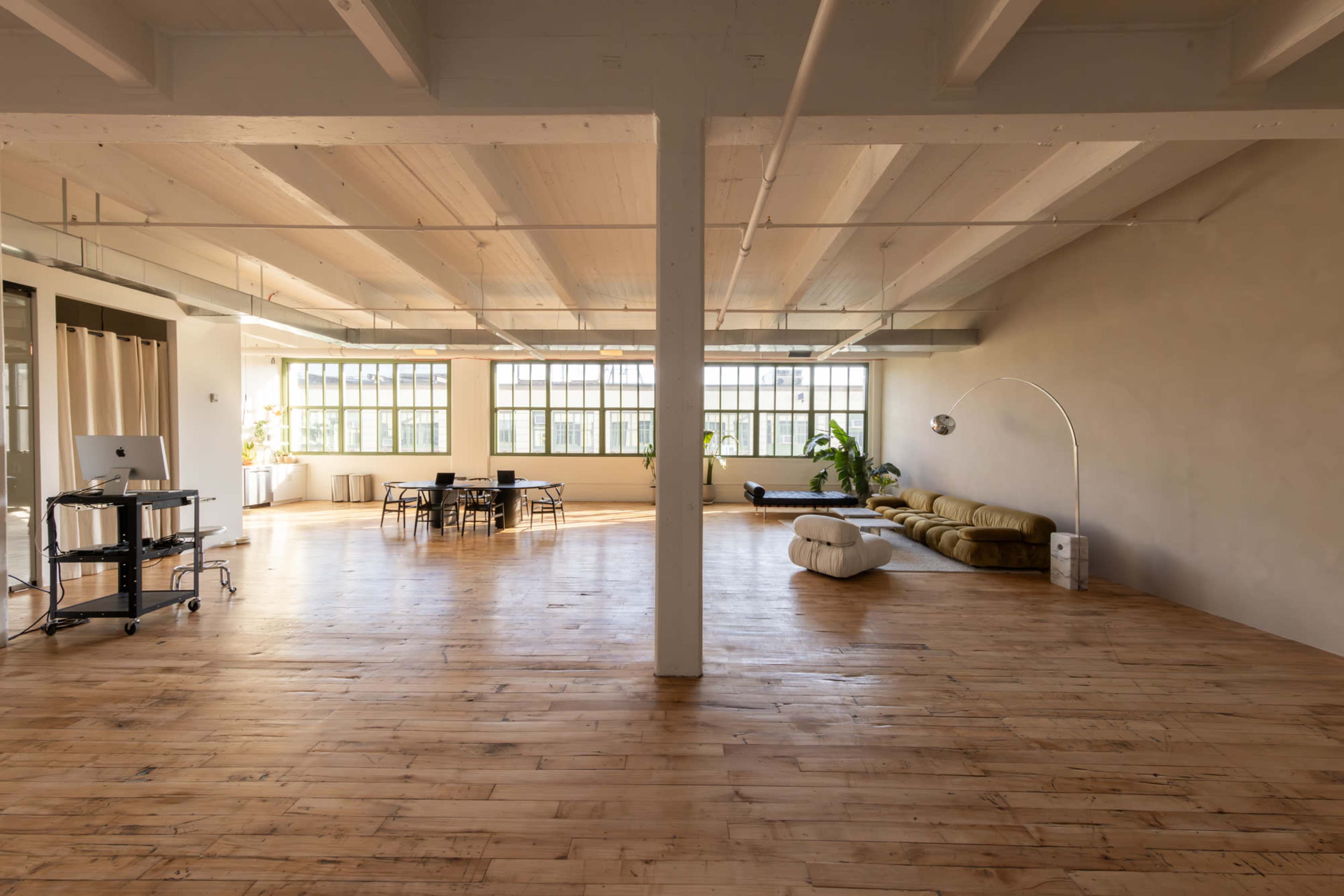 The image shows a spacious, sunlit room with wooden floors, featuring a seating area with a sofa, a table with chairs, and large windows.