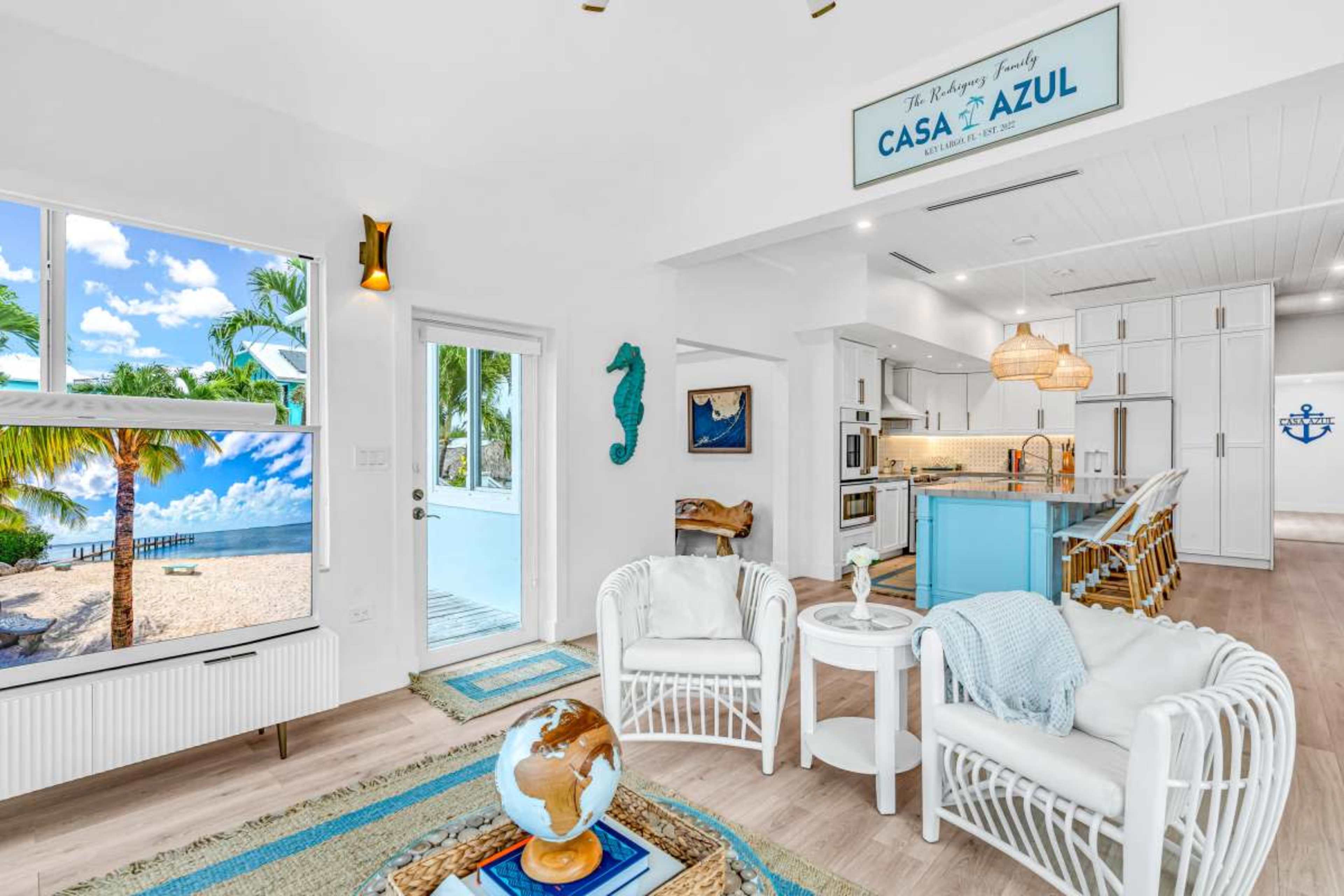 The image shows a bright, modern living space with a beach view, featuring white furniture and light blue accents.