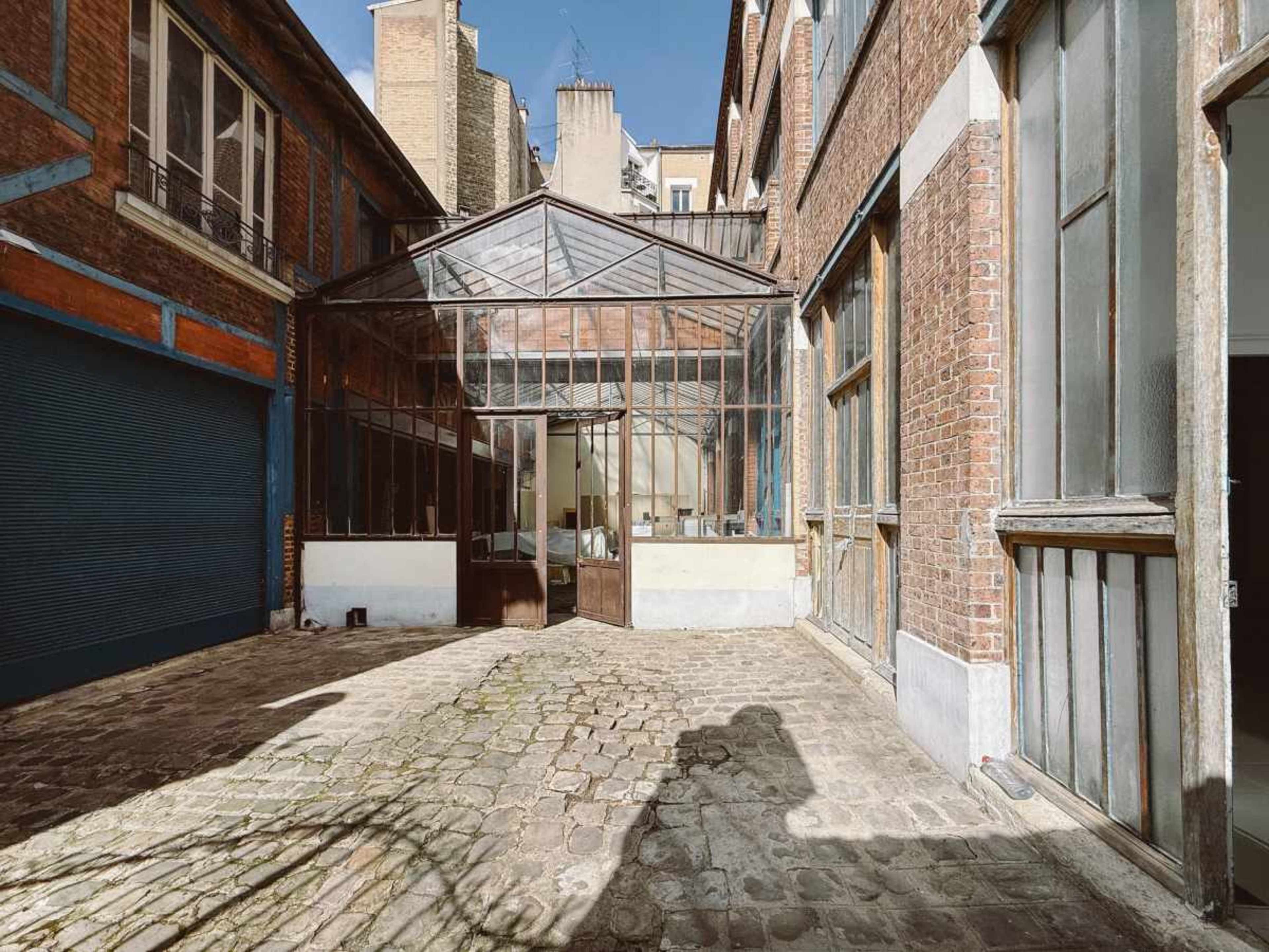 The image shows a cobblestone courtyard with a glass and metal greenhouse structure adjacent to brick buildings.