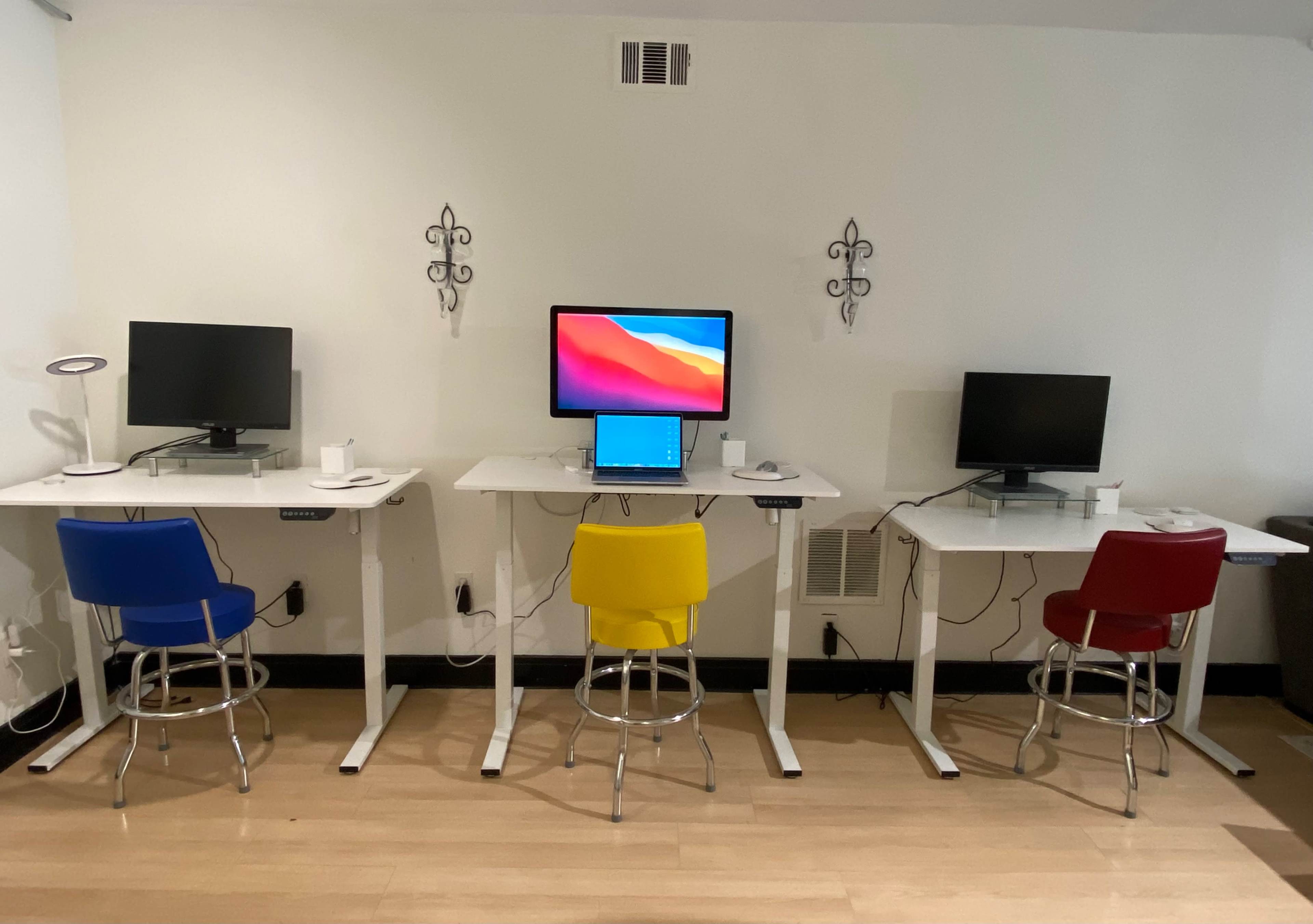 The image shows a room with three standing desks, each equipped with a computer monitor and a laptop, and accompanied by colored bar stools in blue, yellow, and red.