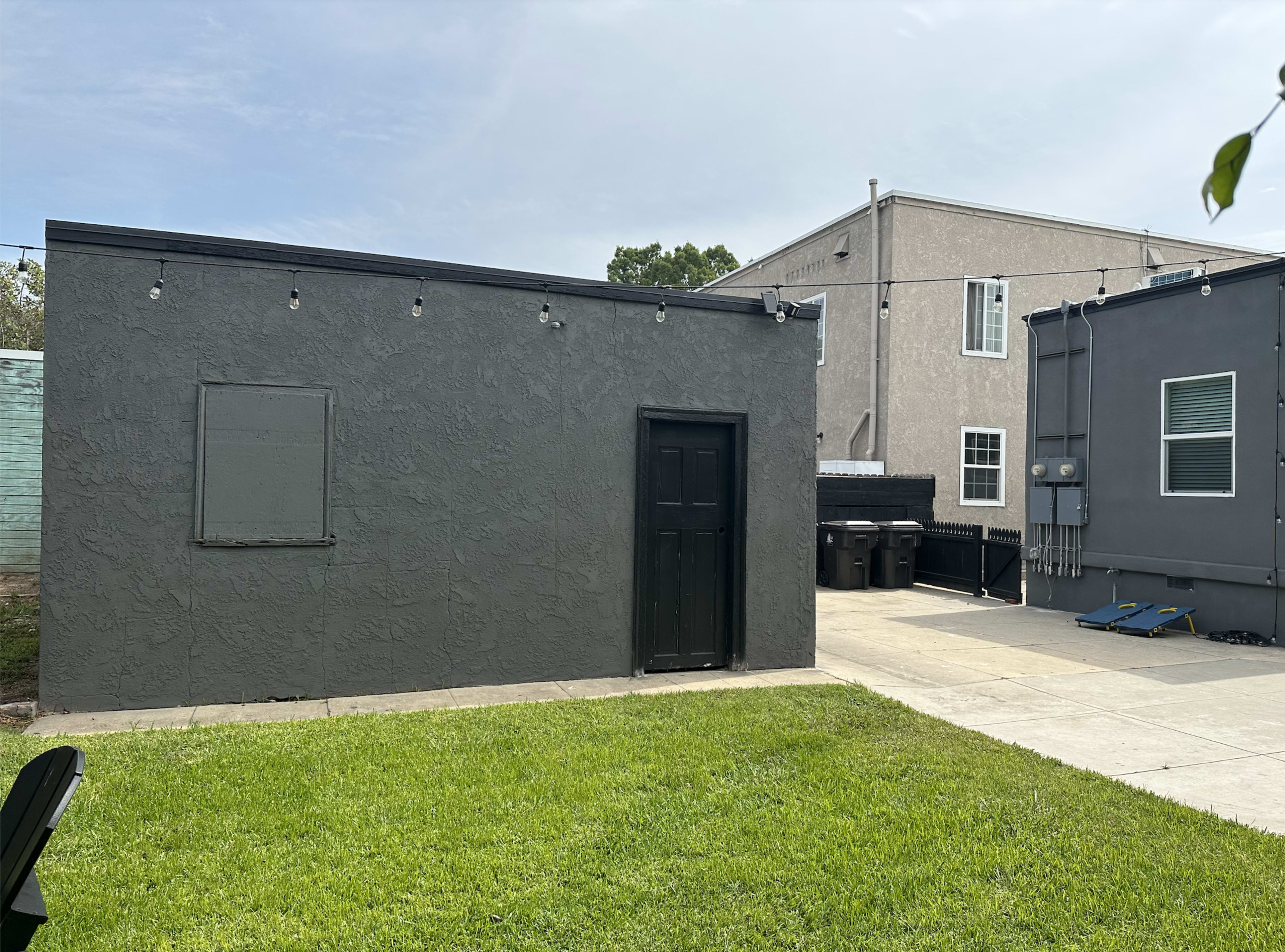 The image shows a gray building with a black door next to a residential structure, along a paved pathway and a grassy area.