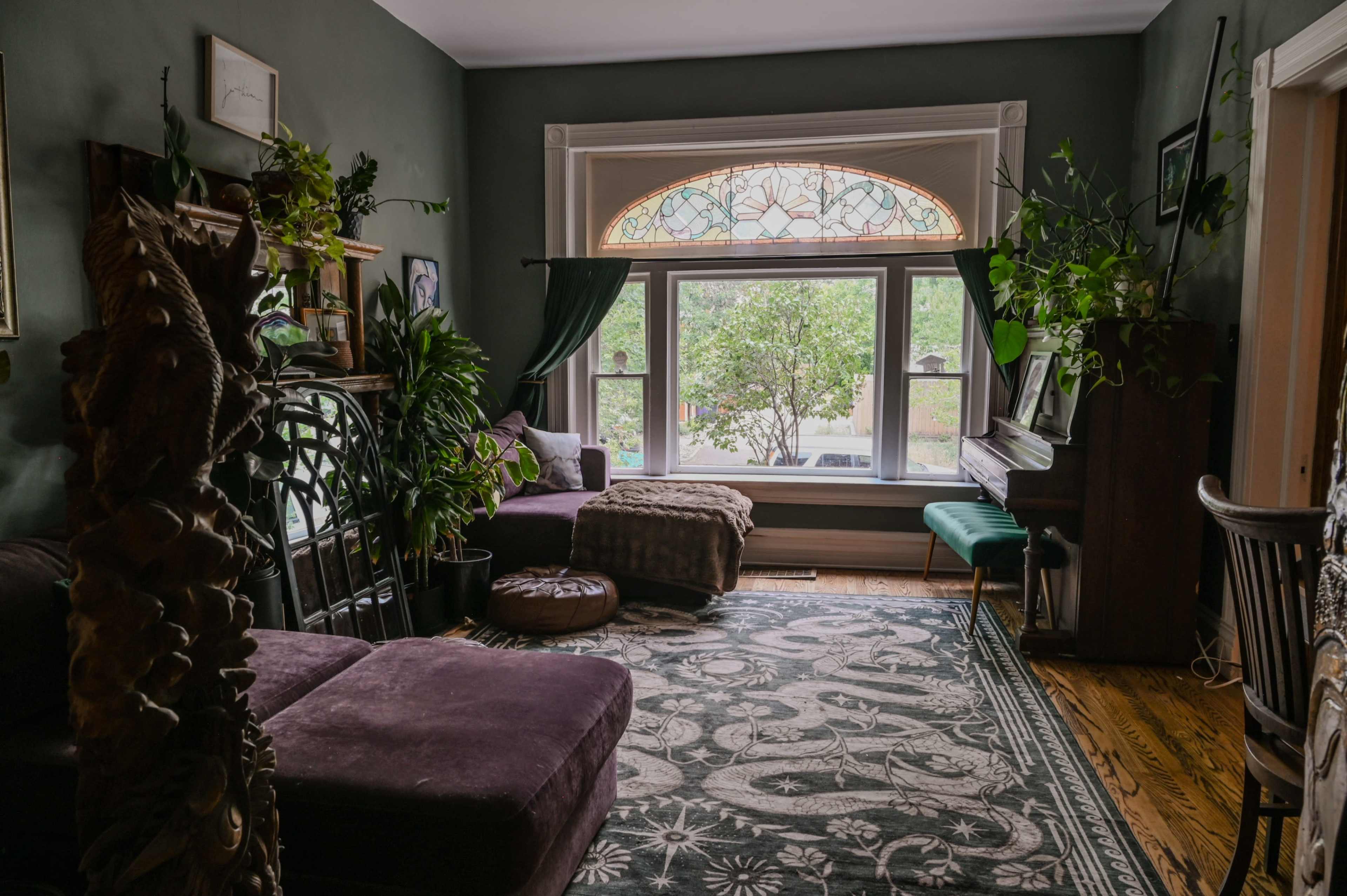 The room features a window with a stained glass design, surrounded by plants, a piano, and a purple sectional couch on a patterned rug.