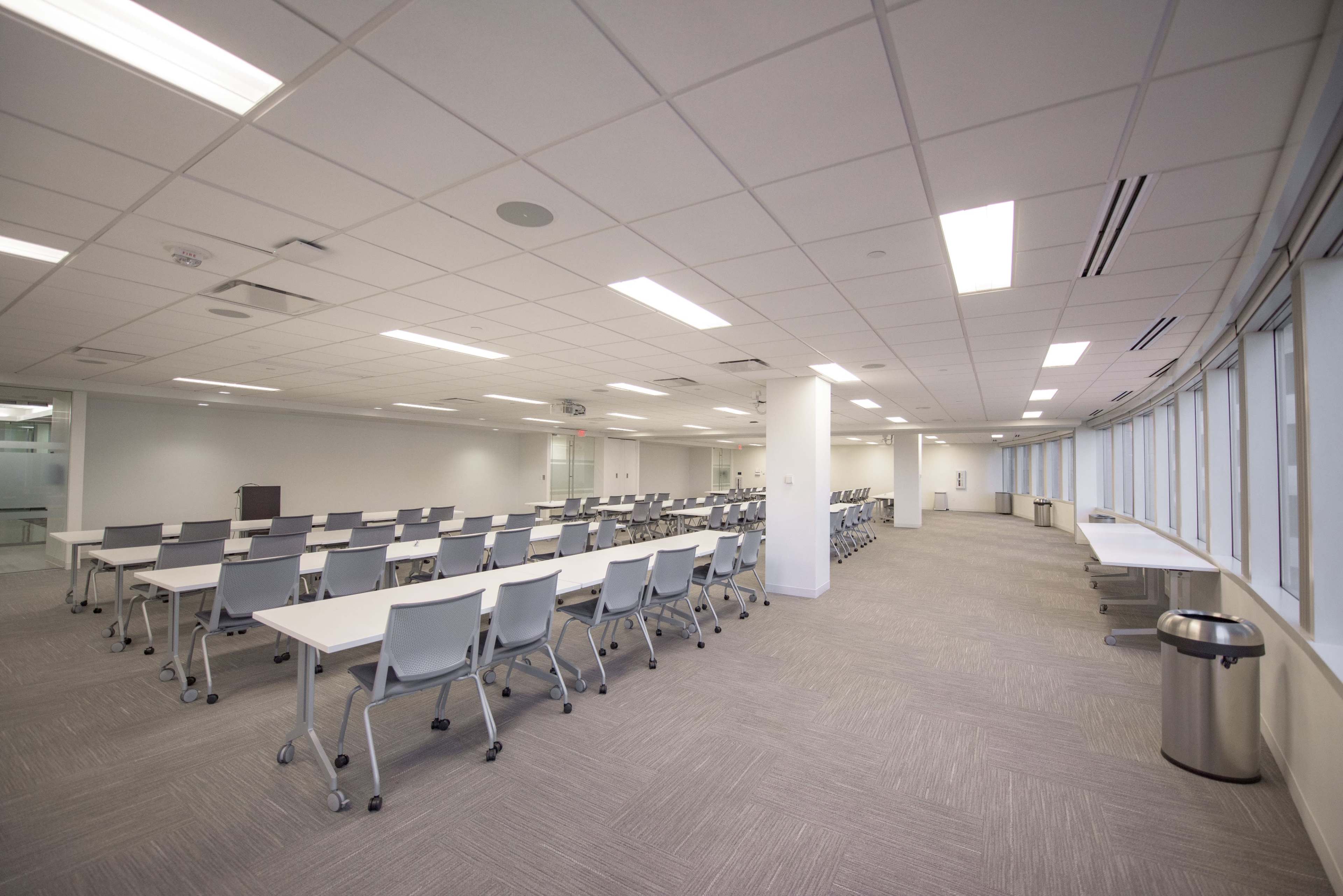 A spacious meeting room features rows of white tables and chairs, with large windows and neutral-colored carpeting.
