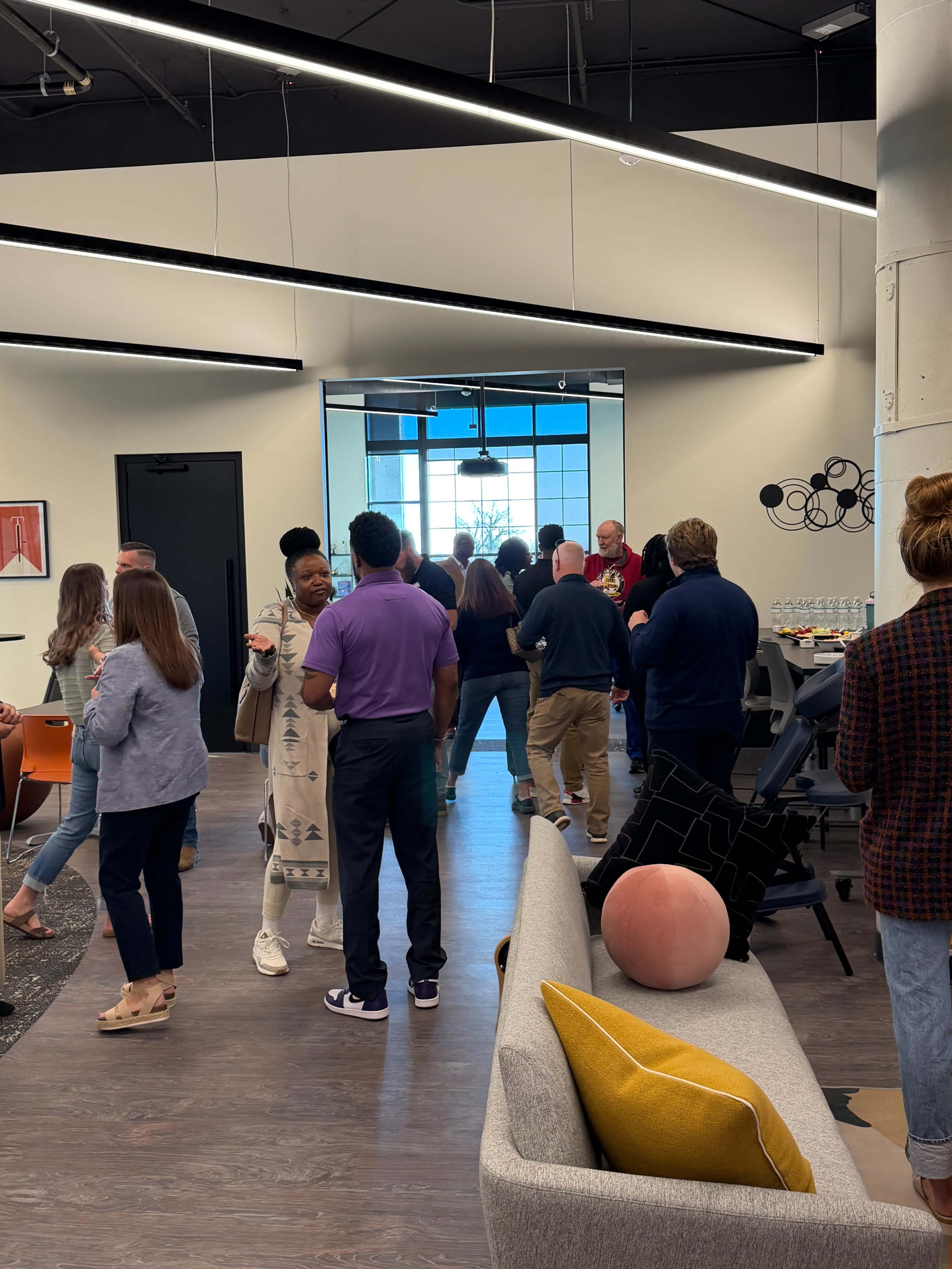 A group of people congregates in a modern, open office space with large windows and contemporary furnishings.