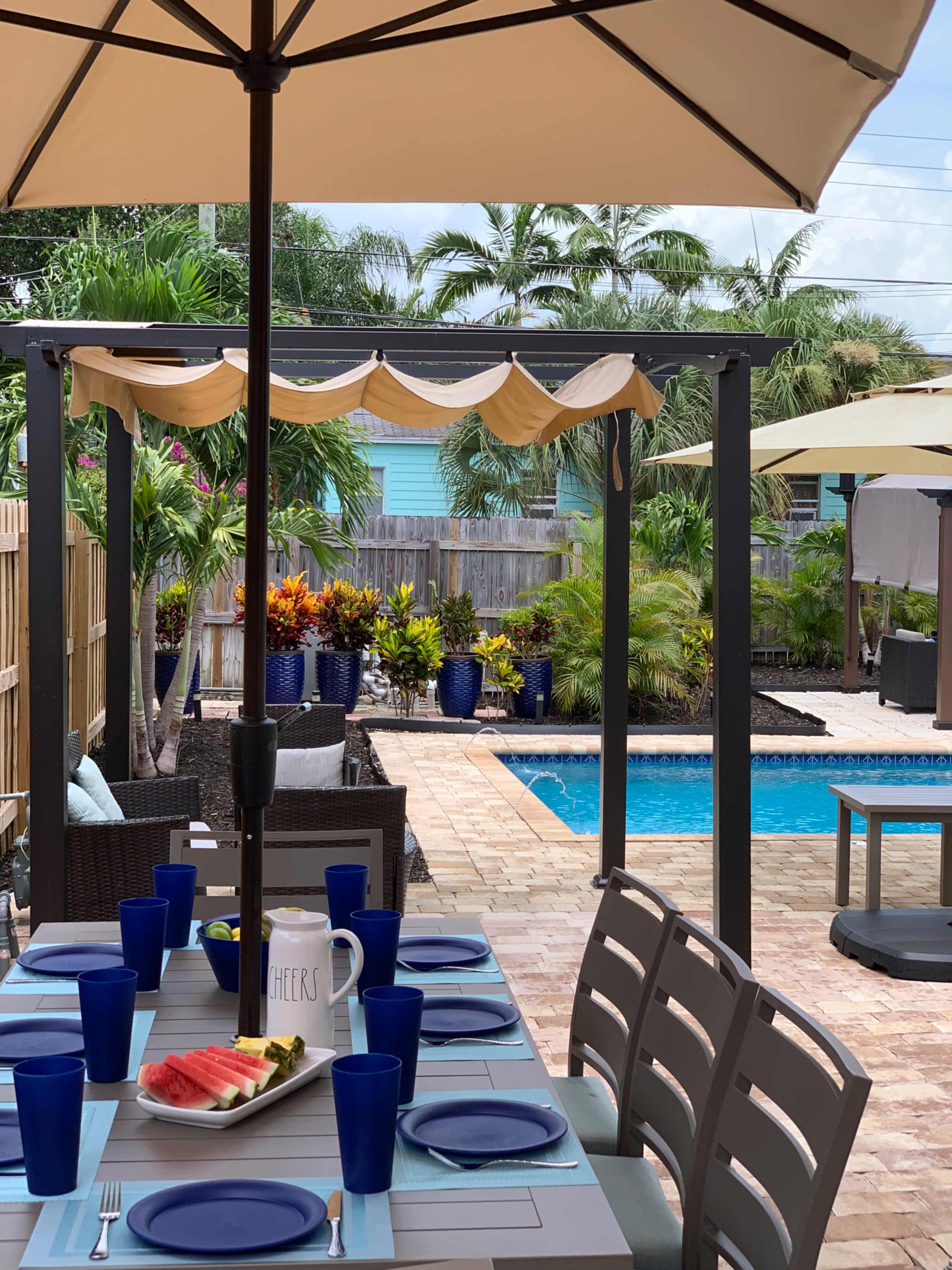 A dining table is set with plates and blue cups near a swimming pool surrounded by tropical plants under an outdoor umbrella.