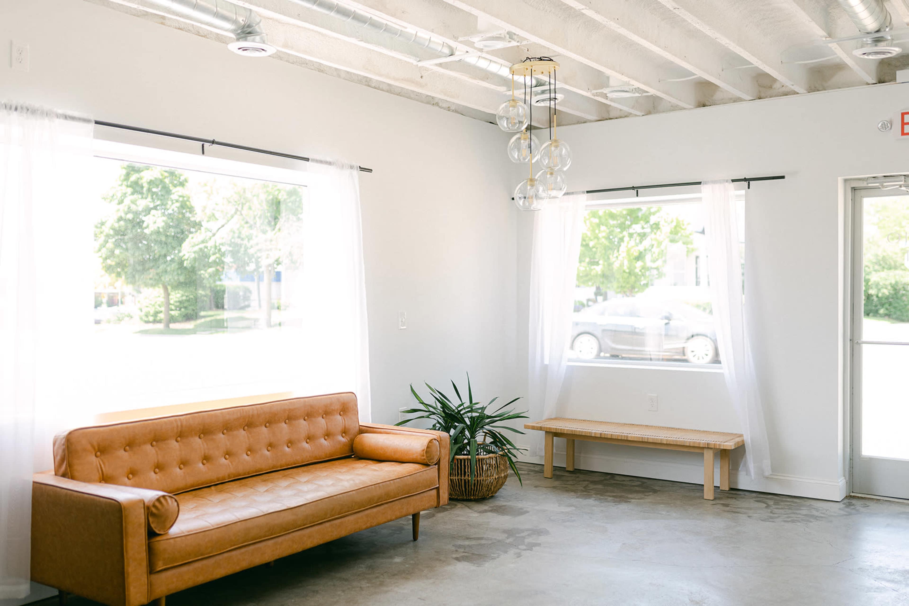 A modern, minimalist room features a brown leather sofa, a wooden bench, large windows with sheer curtains, and a potted plant.