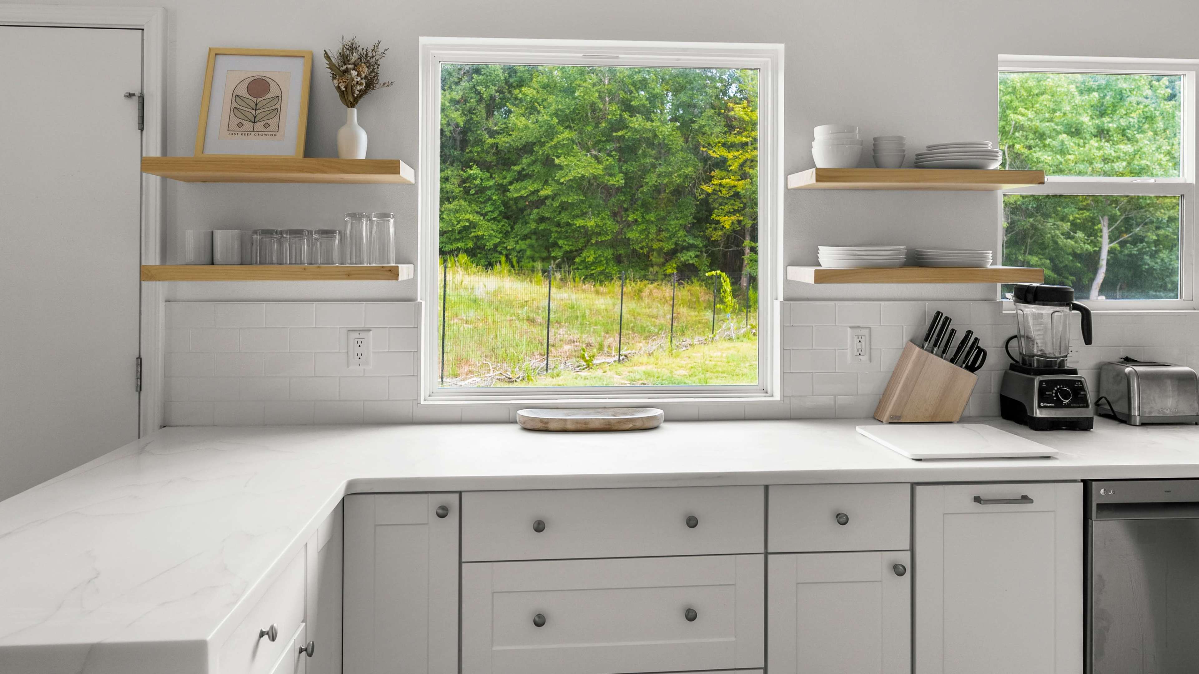 A modern kitchen features a countertop, open shelves with dishware, and a window overlooking a green landscape.