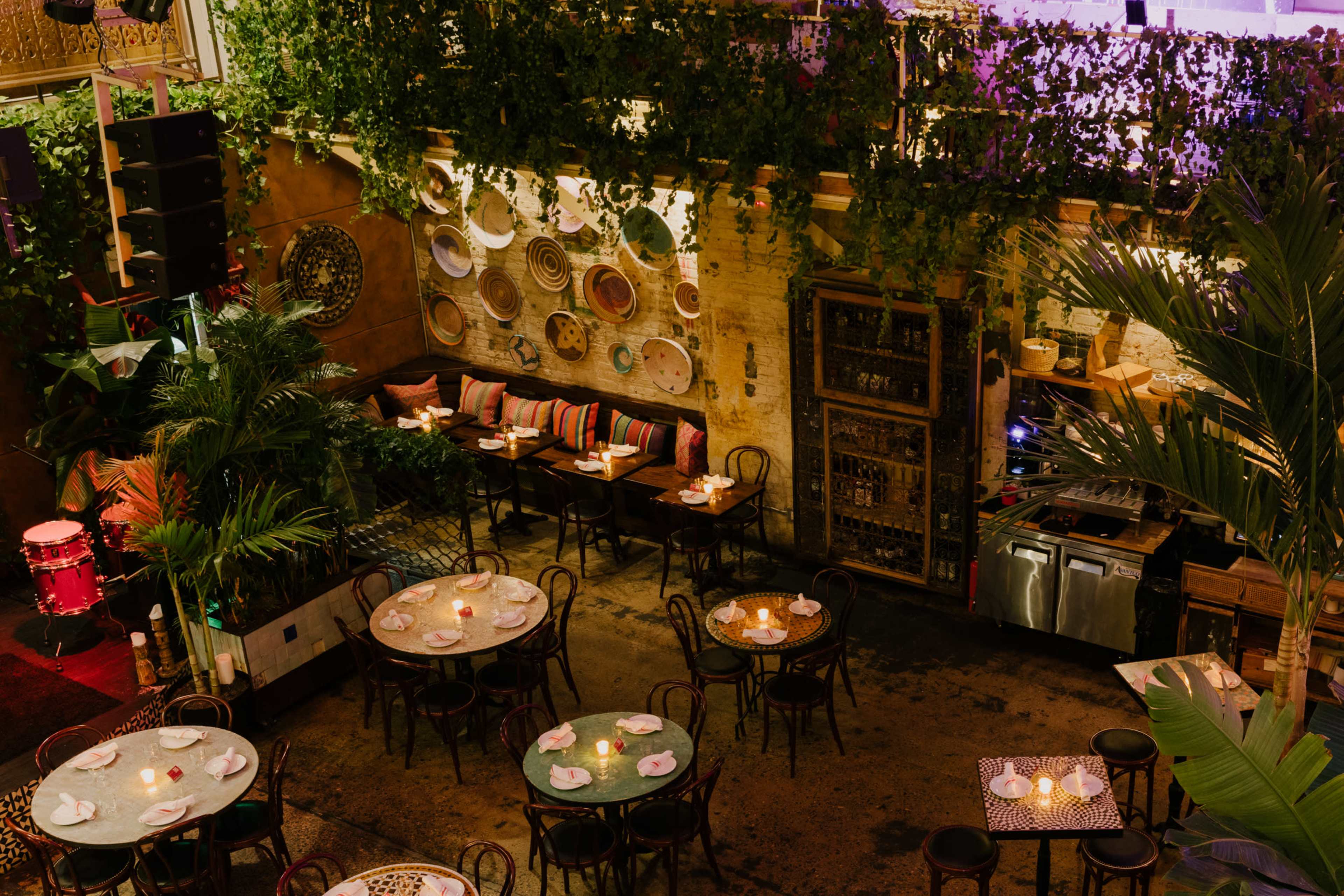 A dimly lit restaurant with several round tables set for dining, surrounded by greenery and decorated walls.