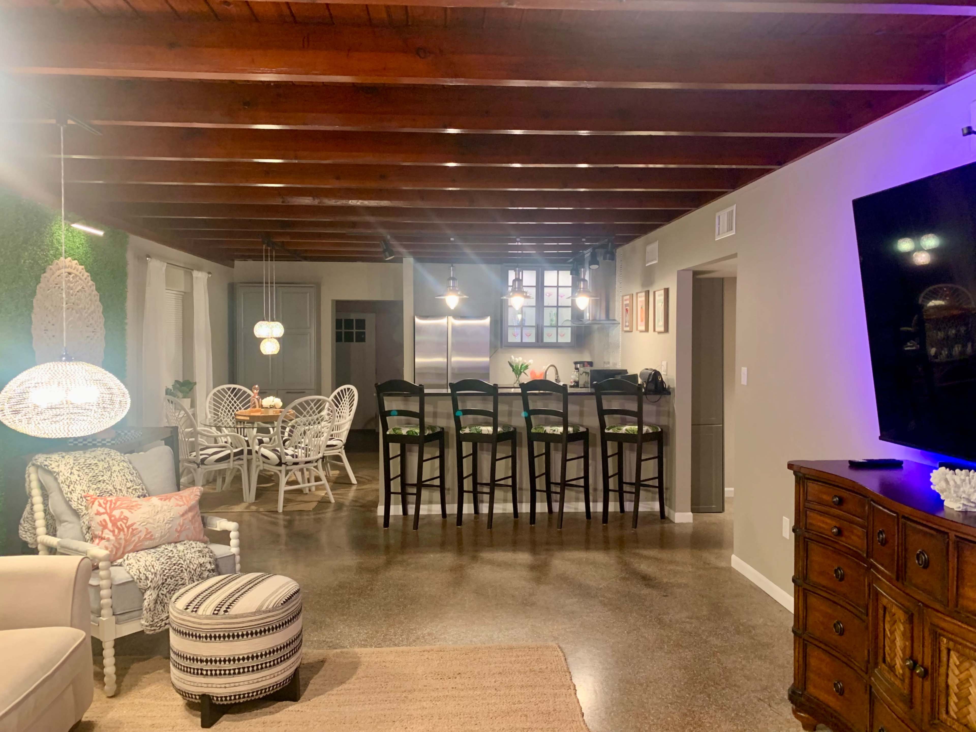The image shows a modern open-concept living and dining area with exposed wooden beams, a dining table surrounded by chairs, and a kitchen in the background.
