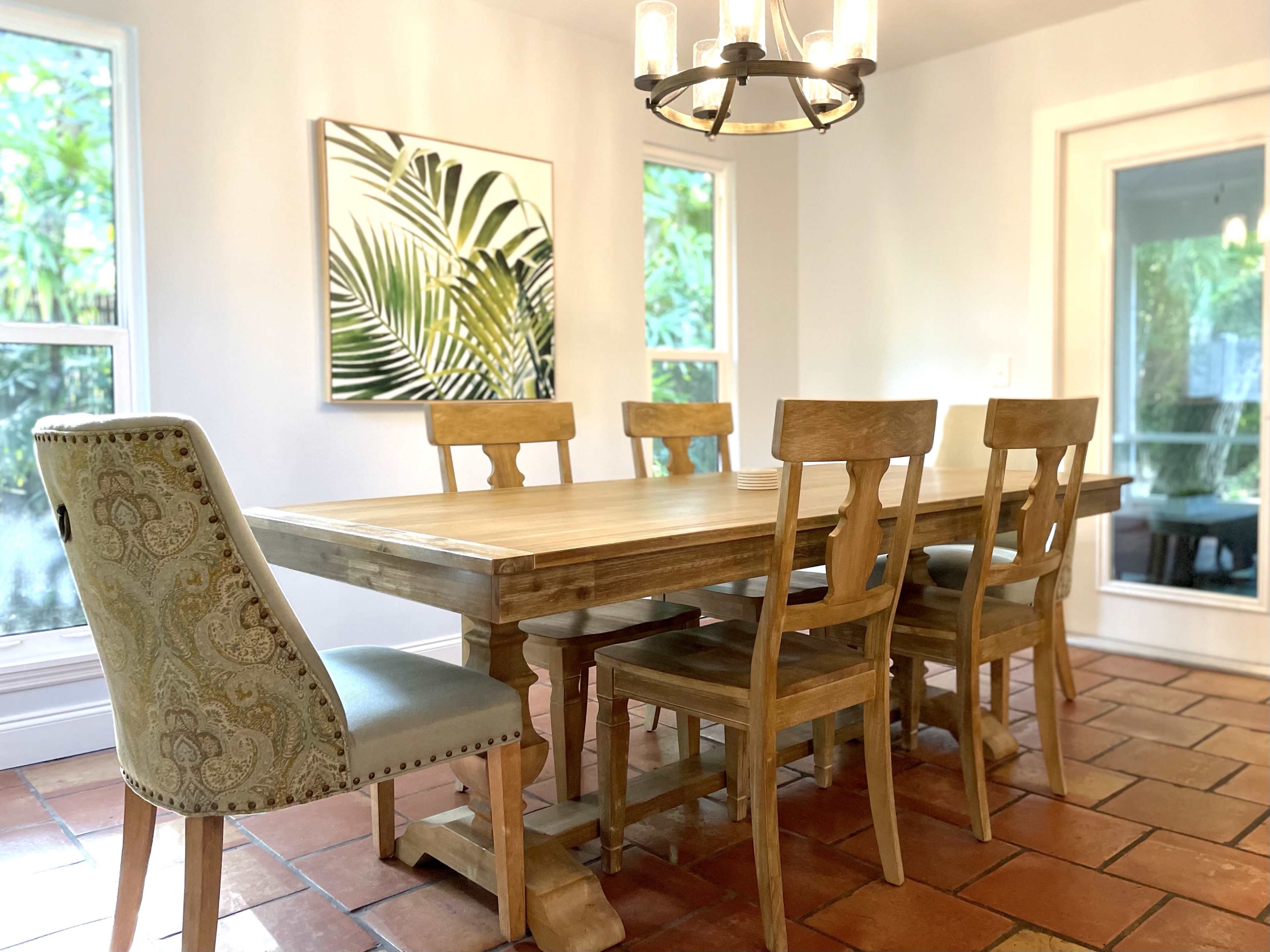 A wooden dining table is surrounded by six chairs in a well-lit room featuring large windows and a tropical-themed artwork on the wall.