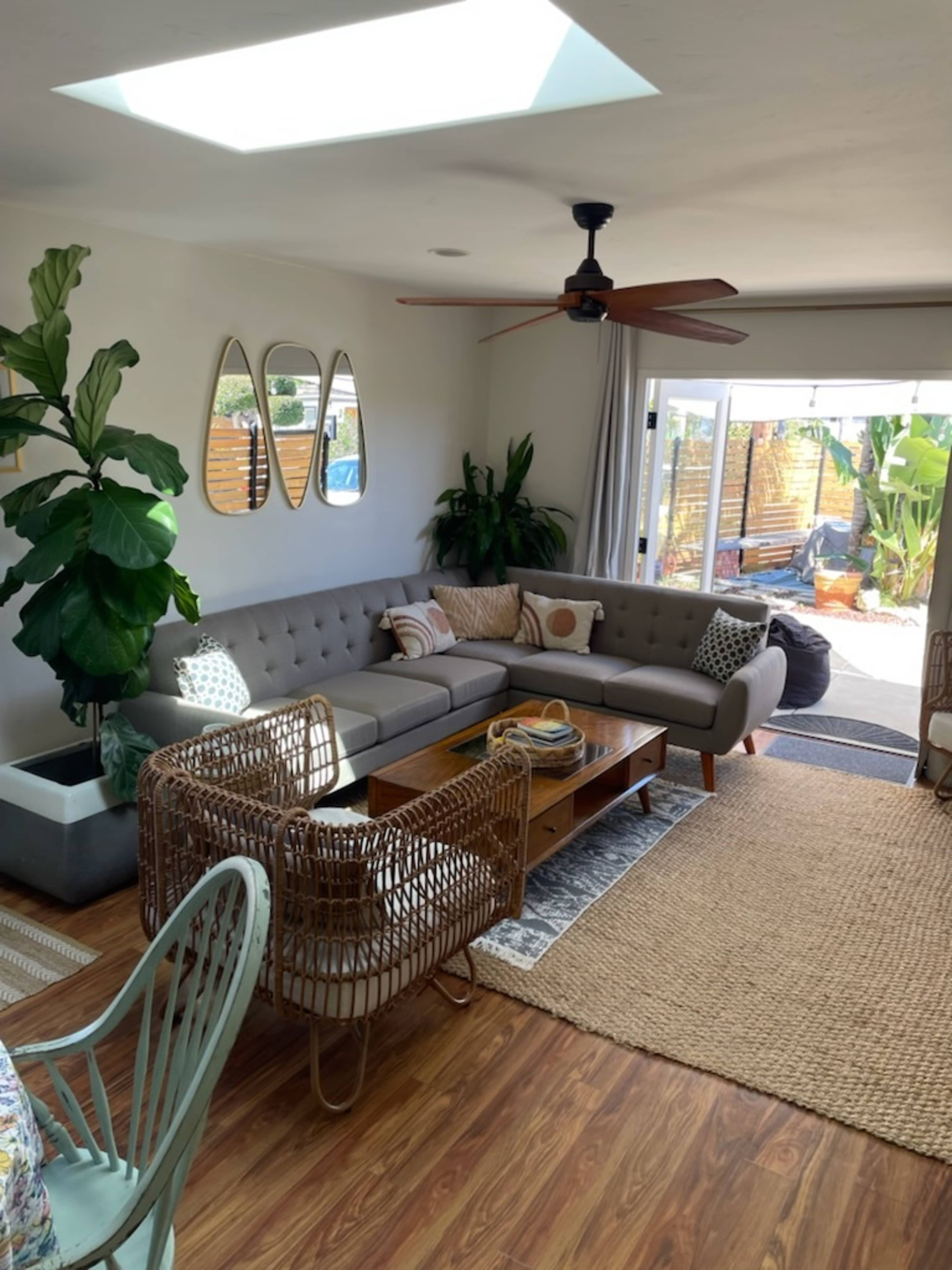 The living room features a gray sectional sofa, a wooden coffee table, and large windows that open to a patio with greenery.