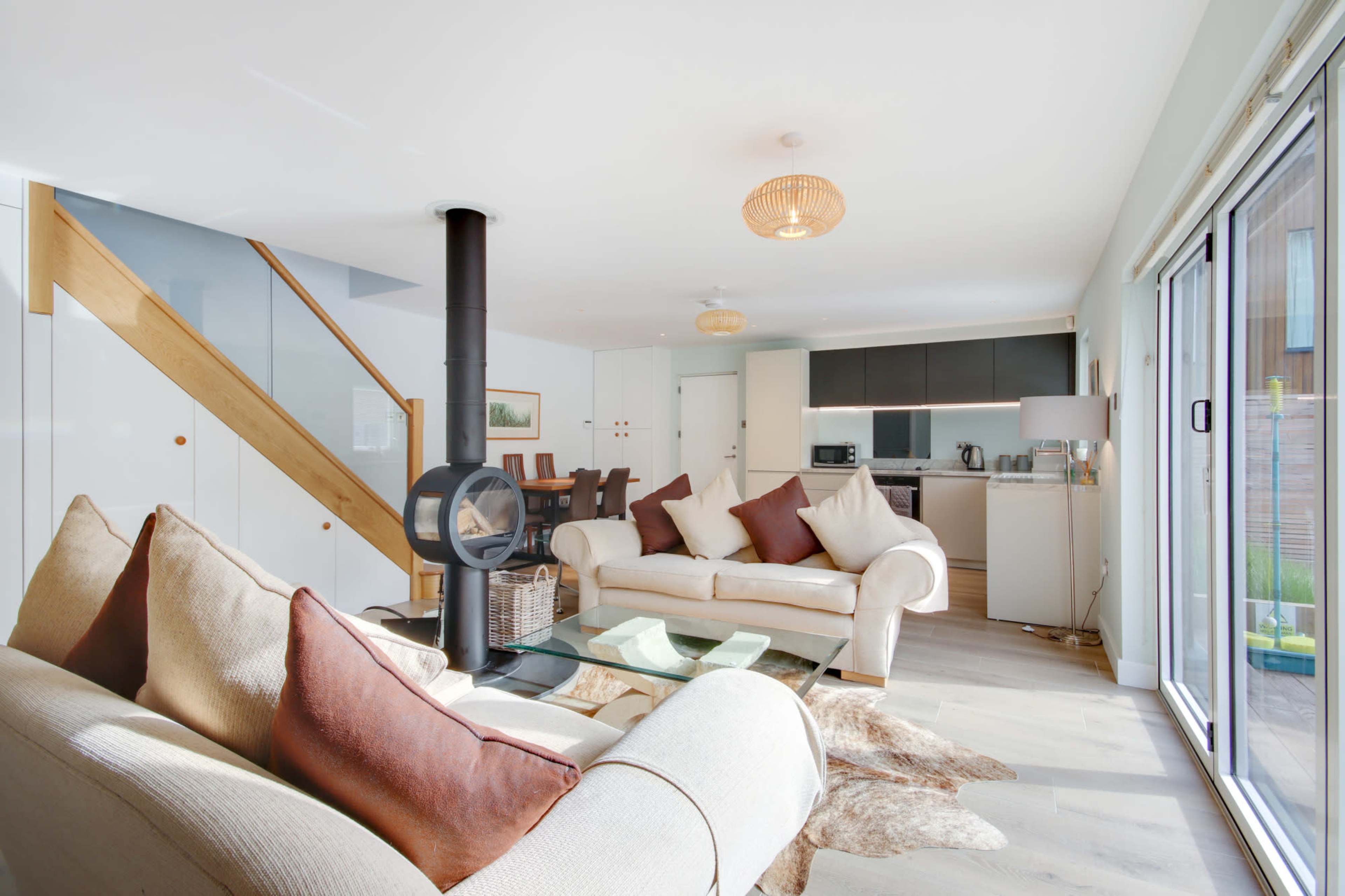 The image shows a modern living area featuring a light-colored sofa with cushions, a glass coffee table, and a fireplace, alongside a dining area with a wooden table in a spacious, well-lit room.