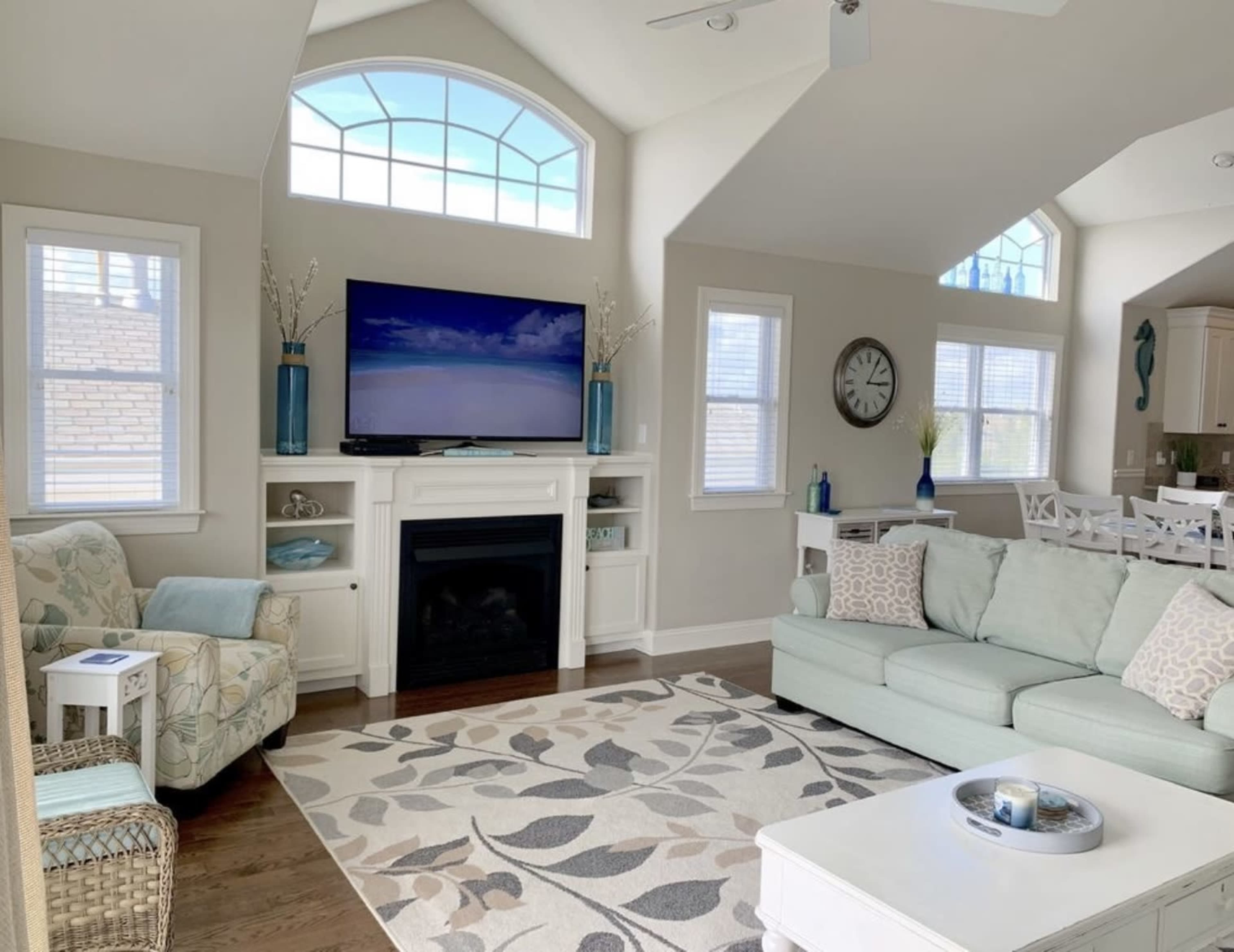 The living room features a large flat-screen TV above a white fireplace, surrounded by light-colored furniture and a decorative area rug with a leaf pattern.