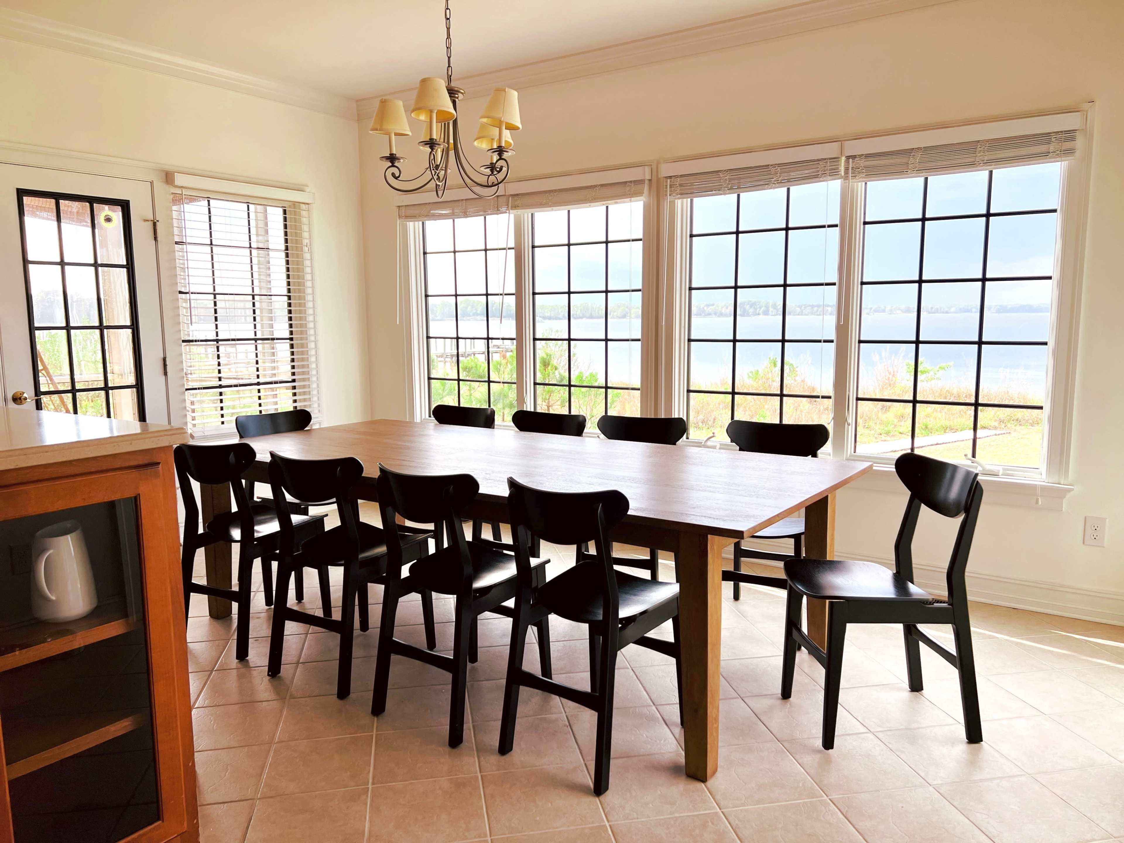 A large wooden dining table with ten black chairs is set in a brightly lit room with multiple windows overlooking a water view.