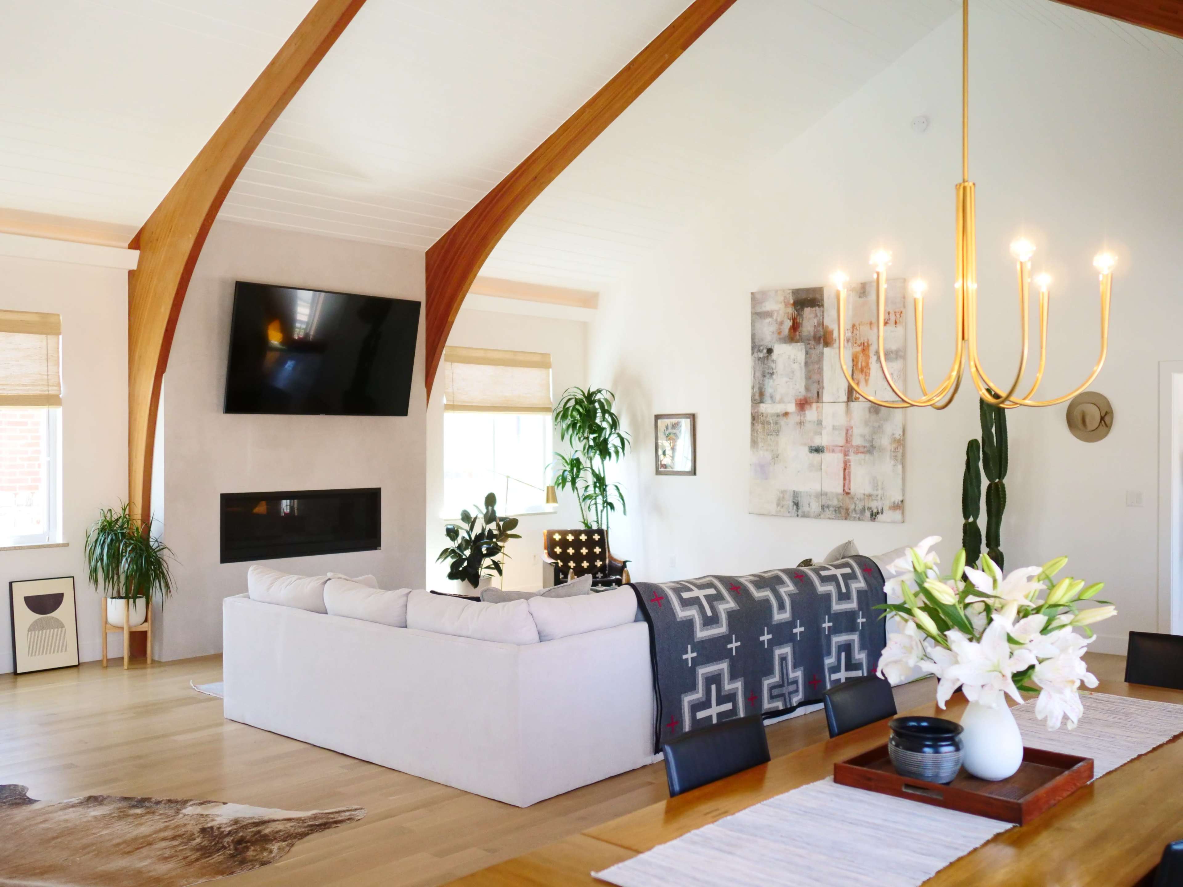 The image shows a modern living room featuring a large sectional sofa, a wall-mounted television, and a dining area with a chandelier and floral centerpiece.