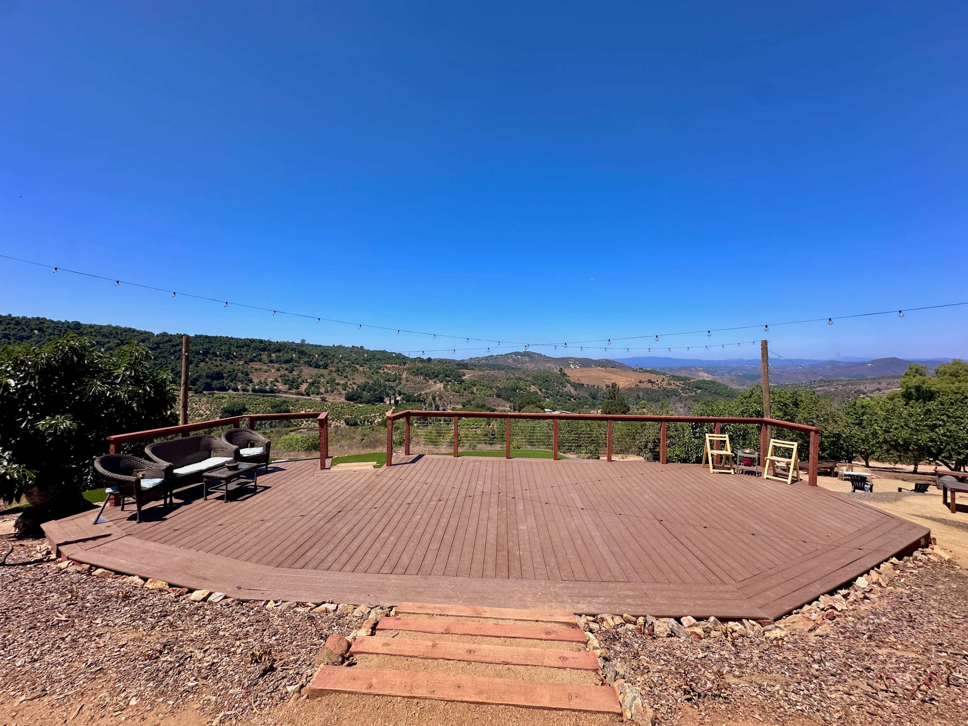 An empty wooden deck overlooks a lush valley under a clear blue sky, with string lights hanging above.