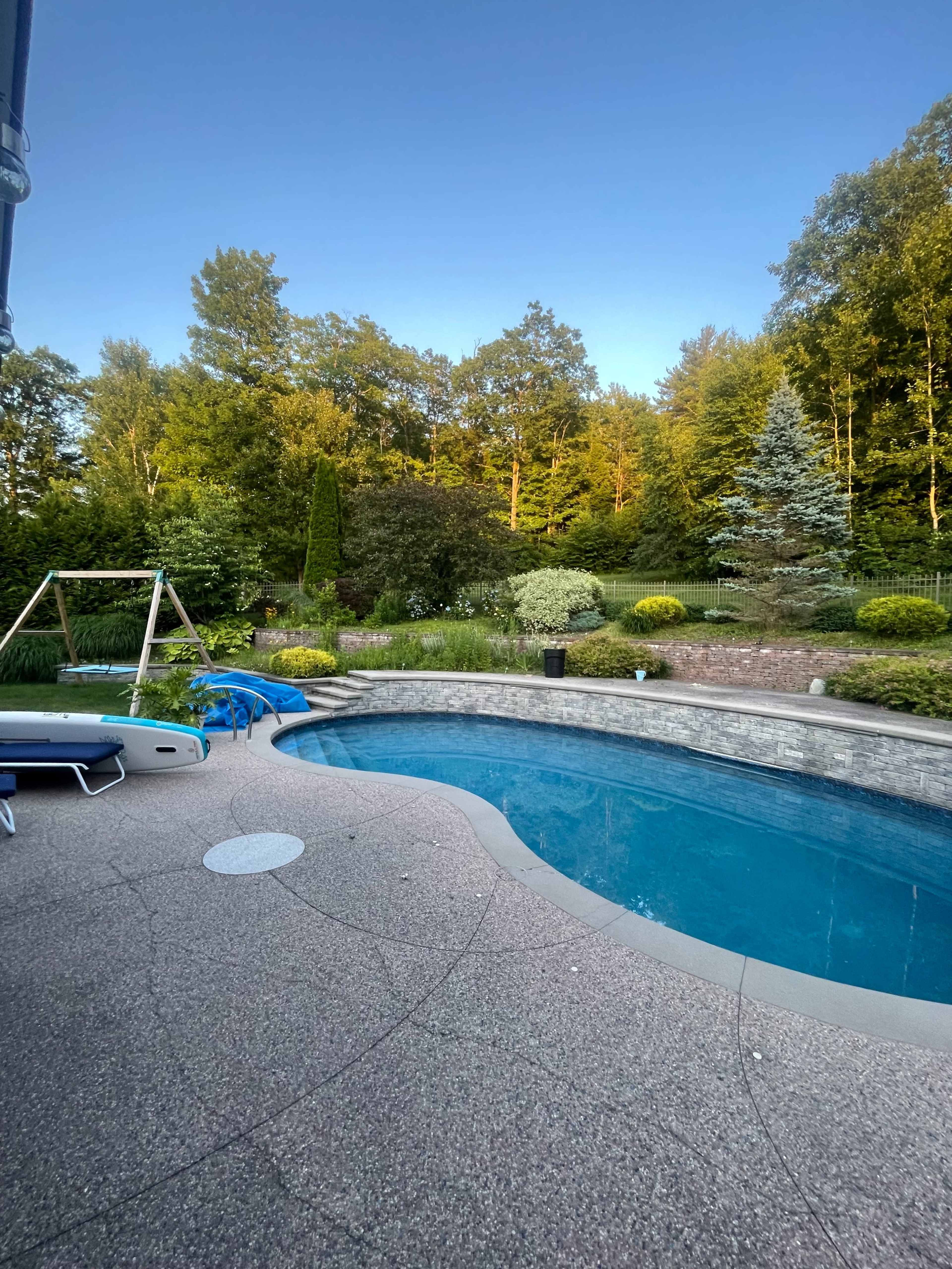 The image shows a backyard scene featuring a swimming pool surrounded by landscaped greenery, a swing set, and a paved area.