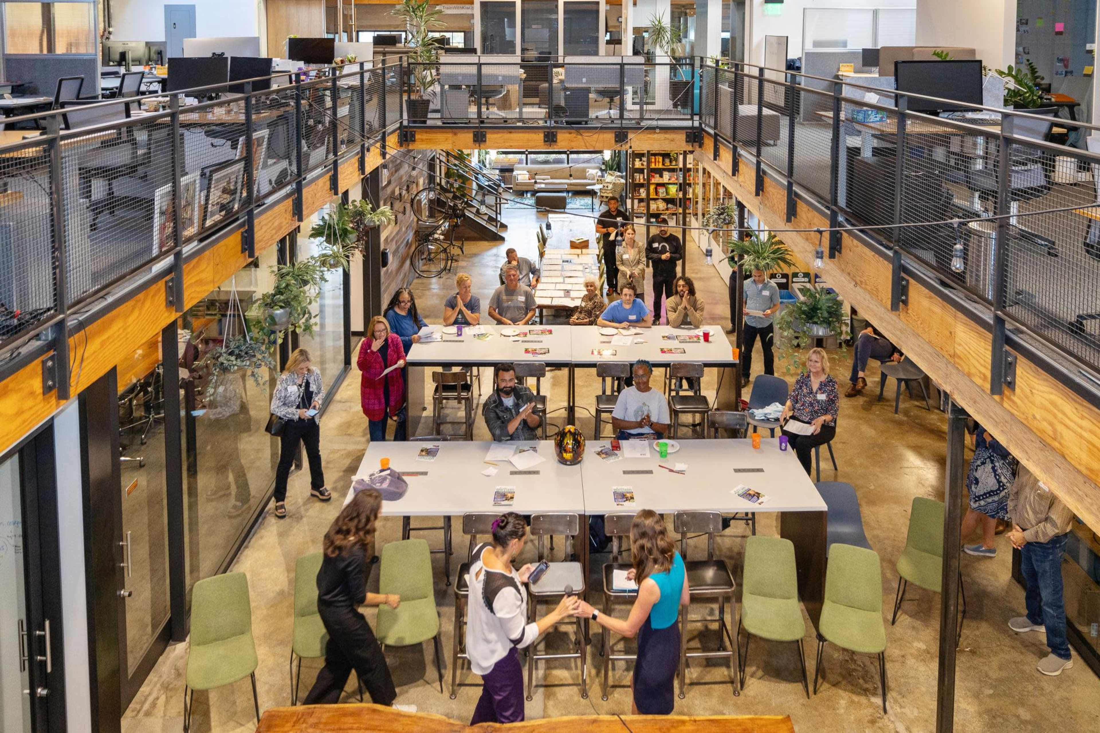 The image shows a spacious open-concept office area with people working and collaborating at tables, surrounded by plants and modern furniture.