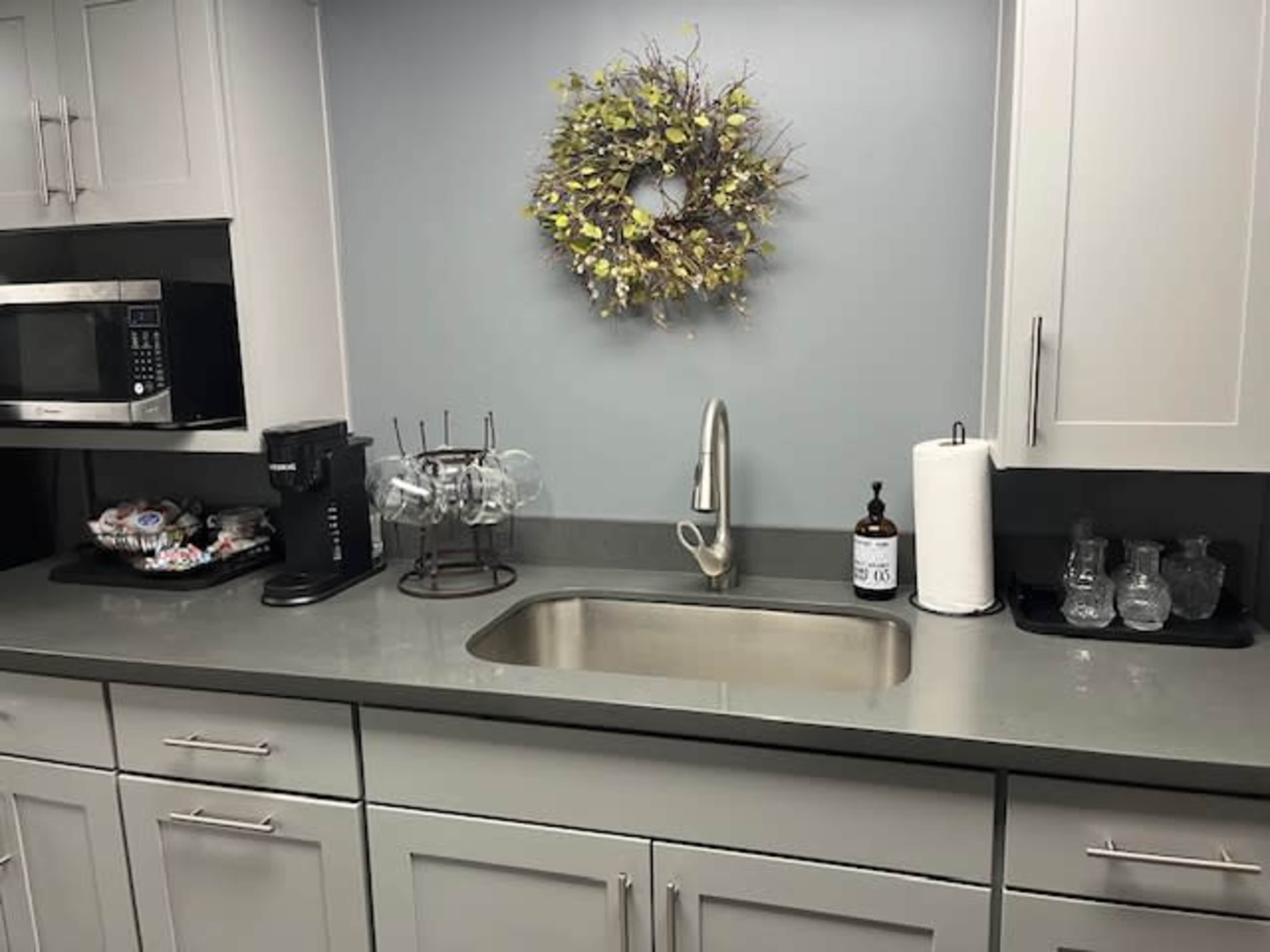 A modern kitchen countertop with a stainless steel sink, a coffee machine, a wall-mounted wreath, and various kitchen supplies organized on the surface.