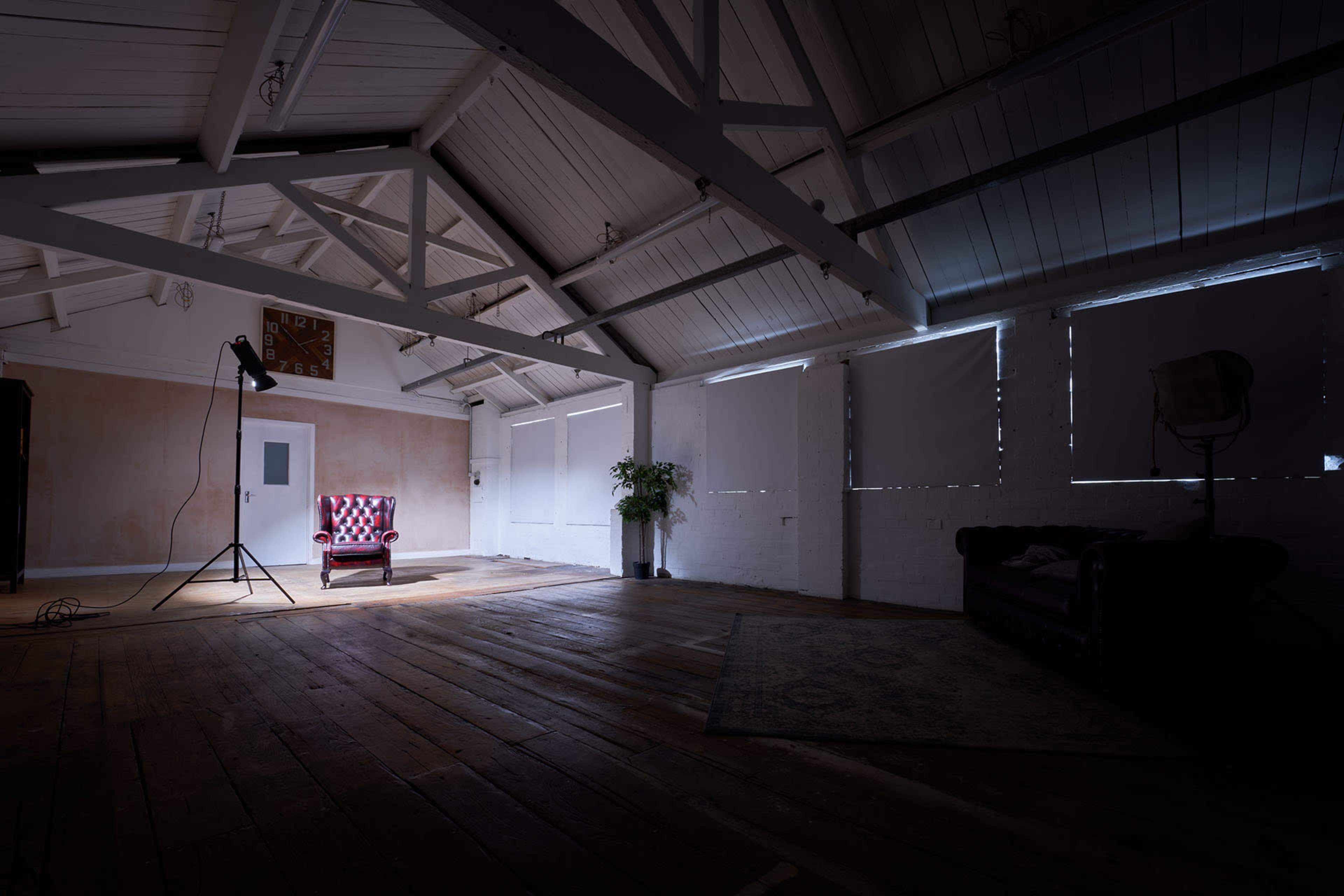 A red and white checked armchair sits in the center of a spacious, dimly lit room with wooden floors and high, exposed beams.