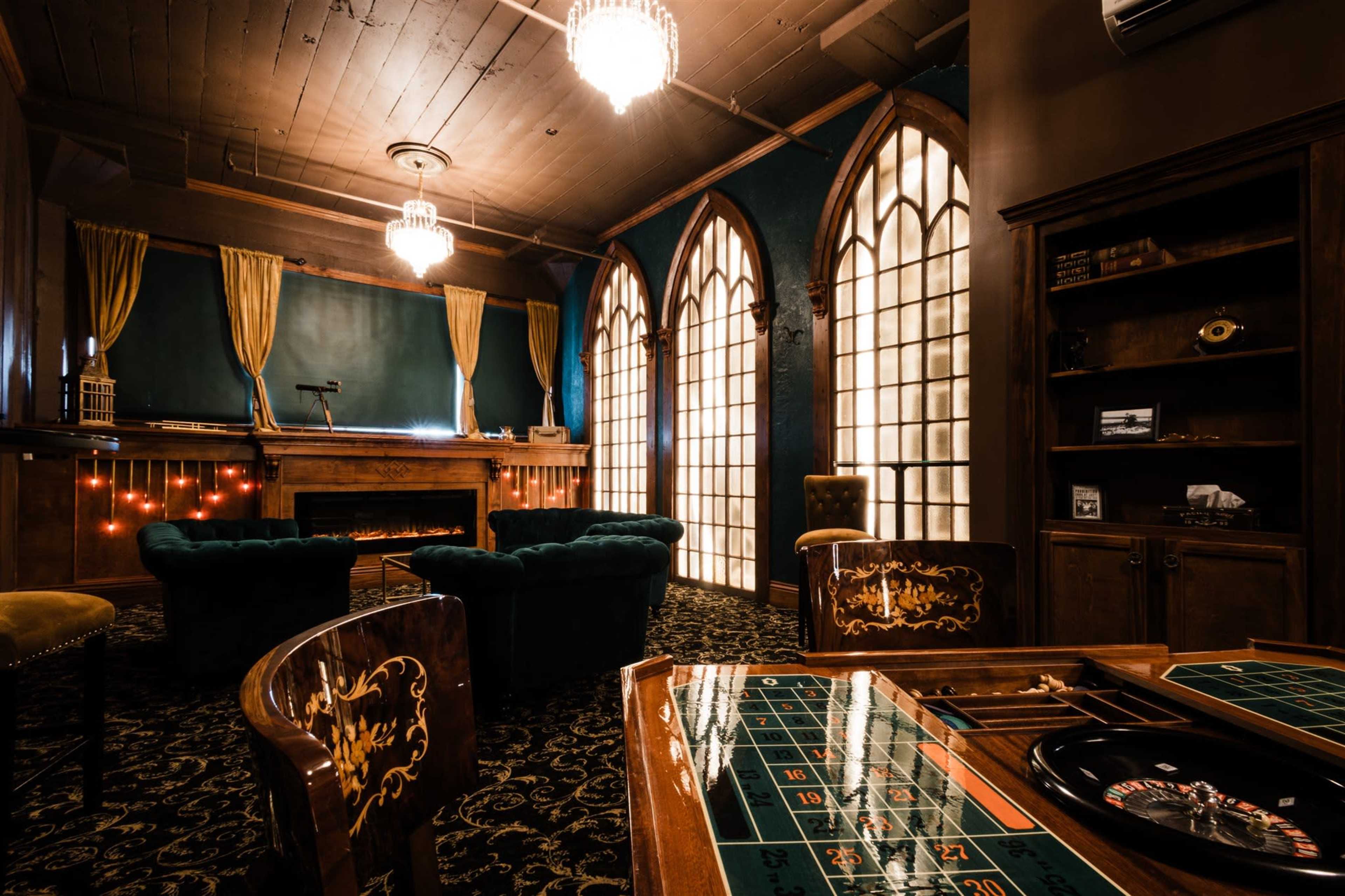 The image shows a stylish, dimly lit room designed for gaming, featuring a roulette table in the foreground and large arched windows in the background.