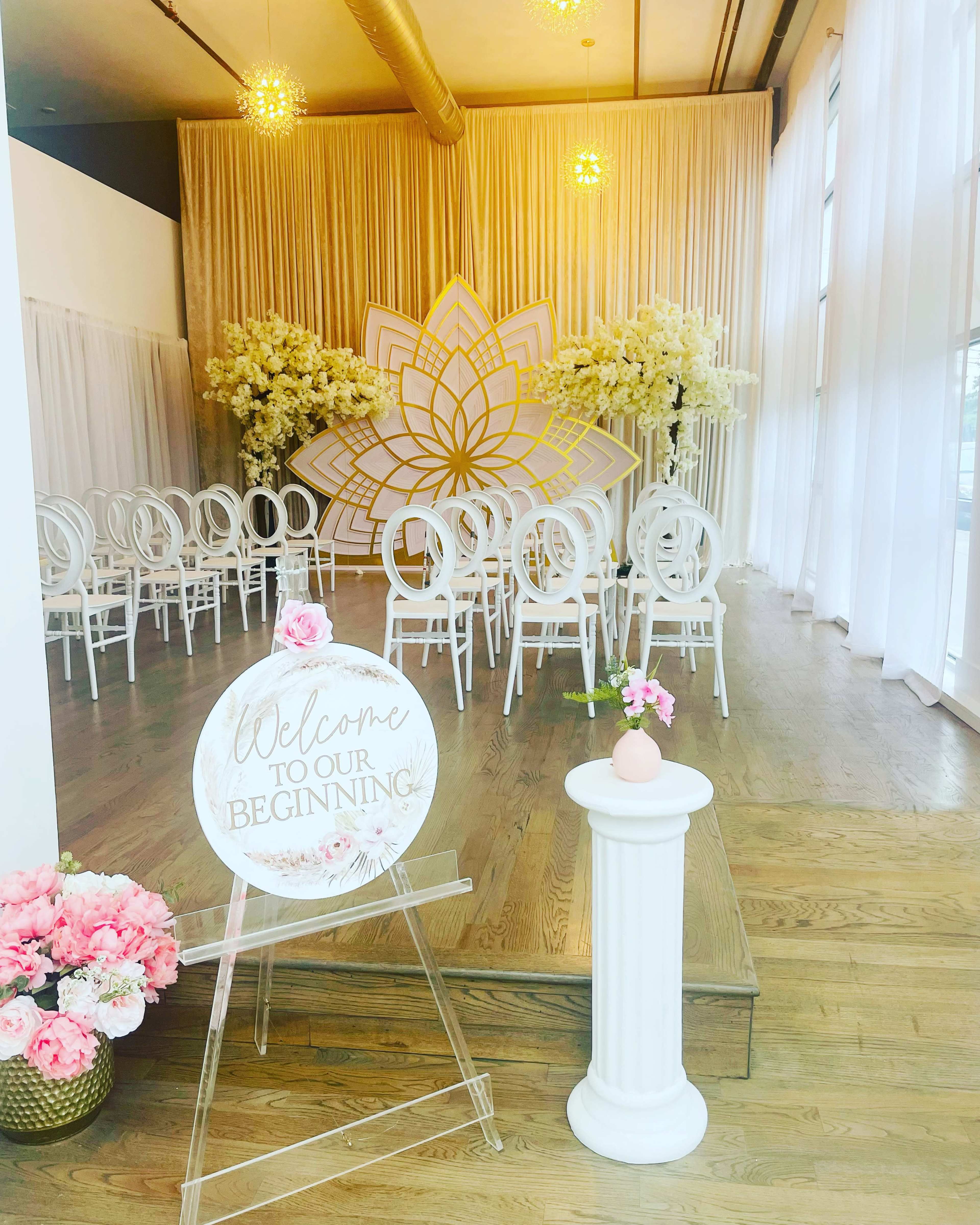 A decorated event space features a large floral backdrop, a circular welcome sign, and rows of white chairs arranged for a ceremony.