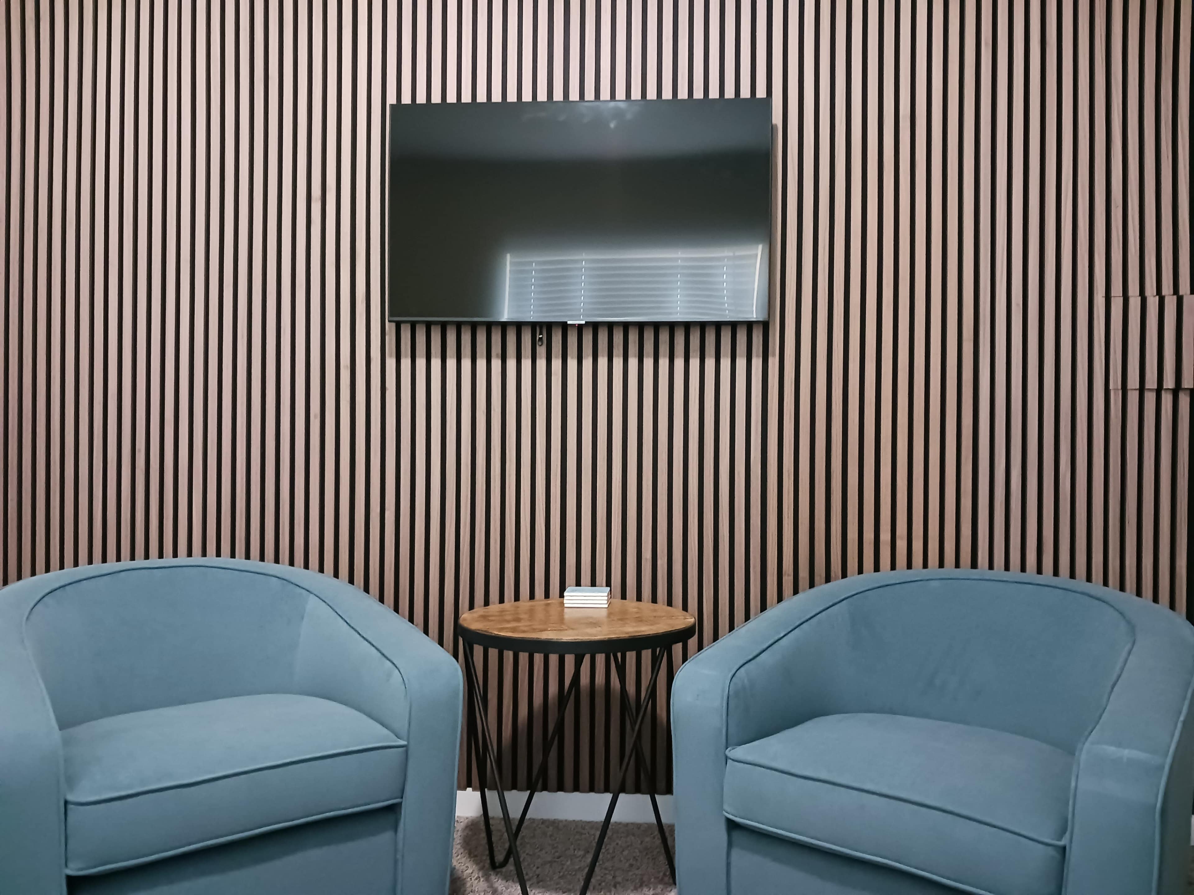 The image shows a seating area with two blue upholstered chairs facing a wooden round table, with a mounted flat-screen TV against a striped wooden wall.