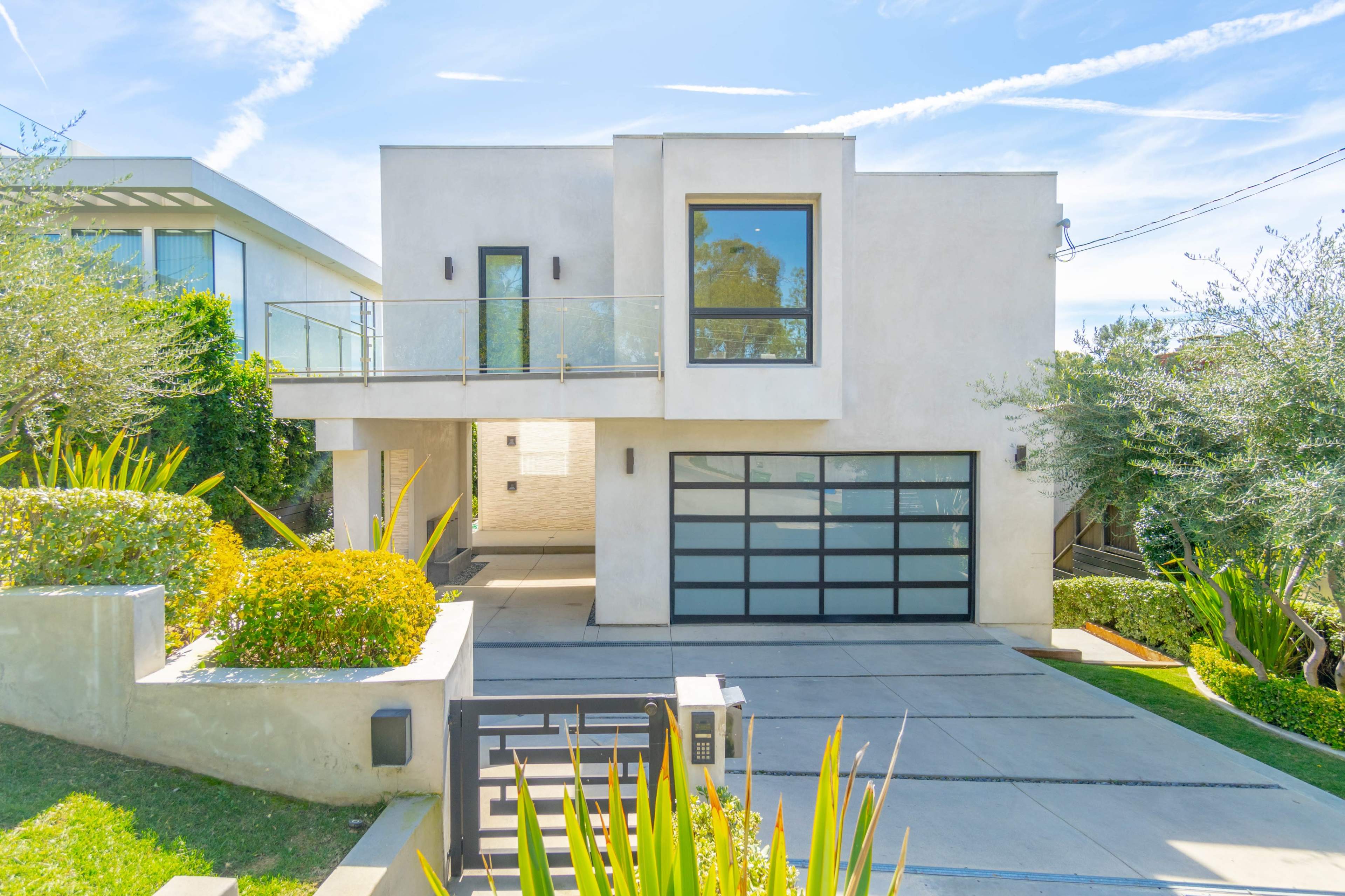 A modern two-story house with a large glass garage door and a balcony, situated on a landscaped lot with greenery.