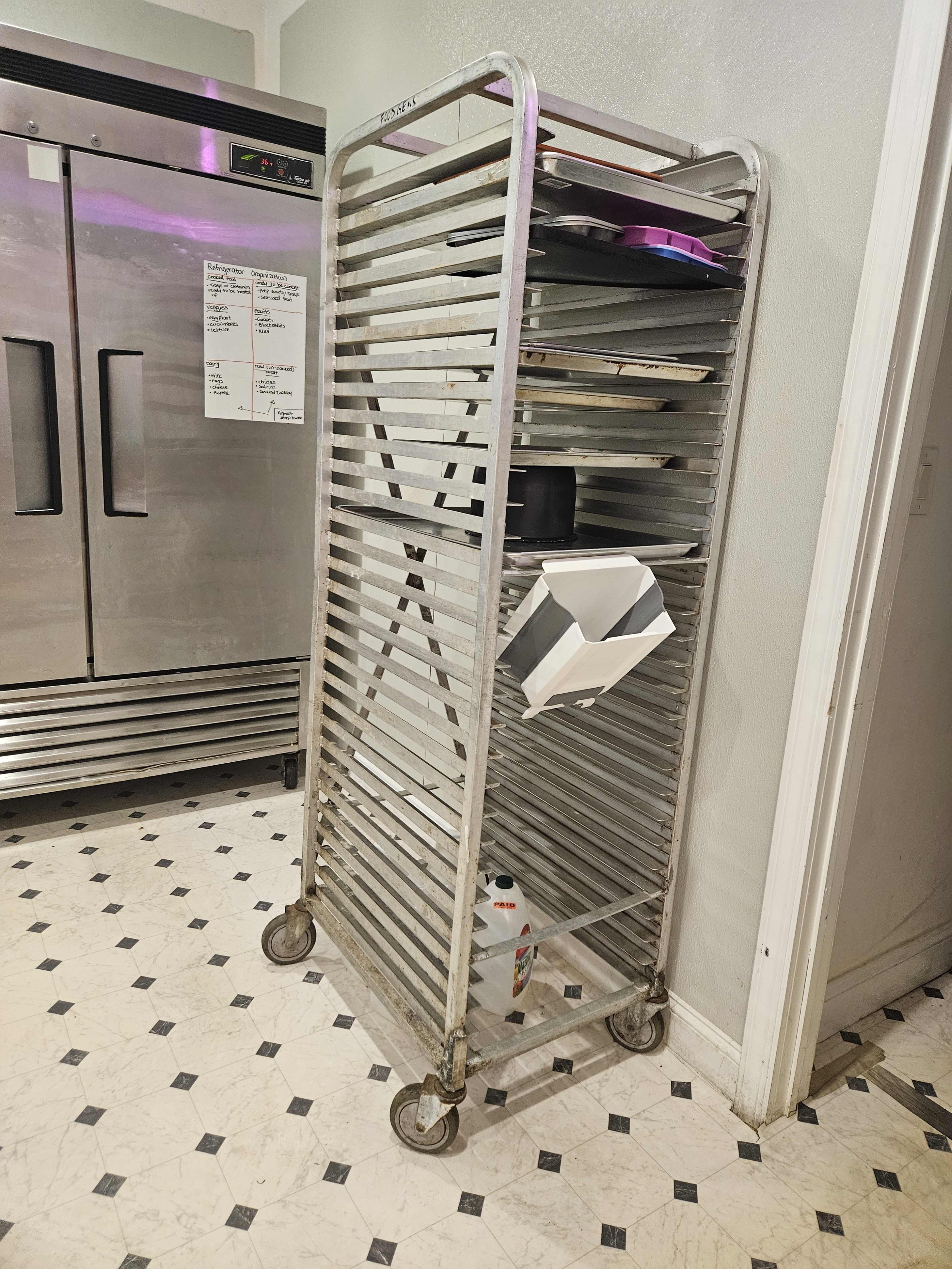 A metal food cart with multiple shelves stands against a wall next to a refrigerator in a kitchen.